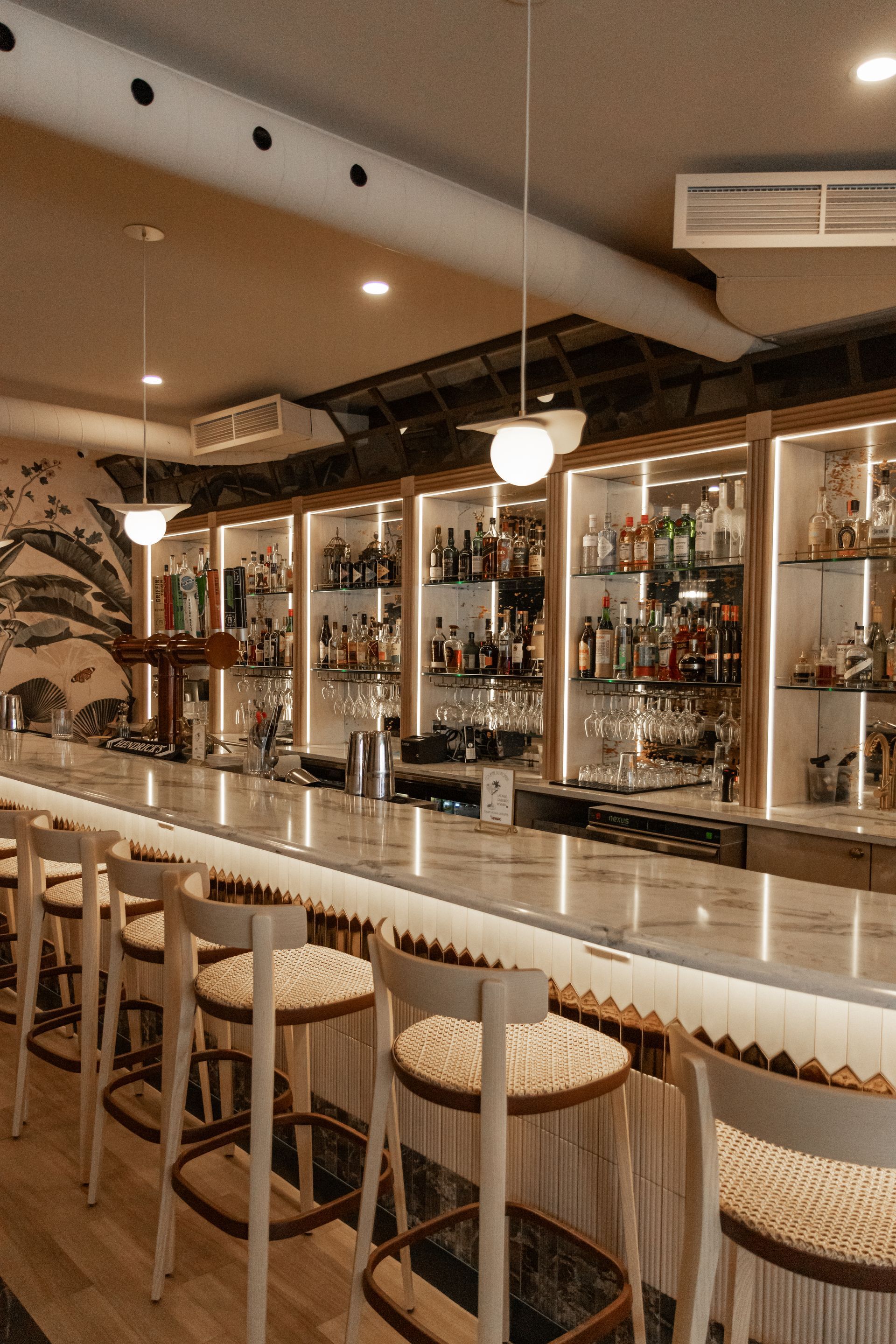 Bar élégant avec étagères à spiritueux rétroéclairées, comptoir blanc et tabourets ronds dans un salon à l'éclairage chaleureux