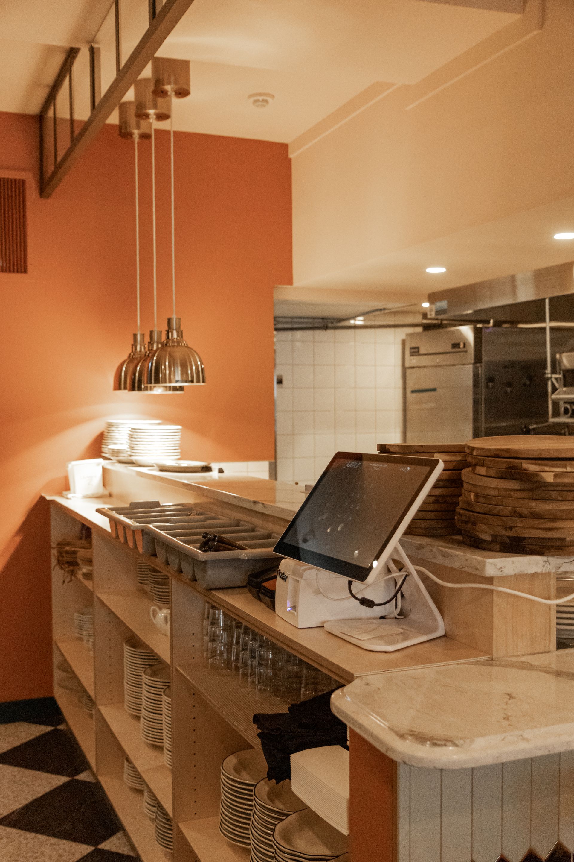 Cuisine moderne avec comptoirs en acier inoxydable, suspensions lumineuses, mur d'accent orange et écran noir sur l'îlot central.