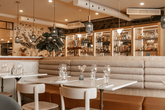 Intérieur de restaurant chaleureux et moderne avec des banquettes beiges, des tables blanches et des étagères en verre derrière le bar.