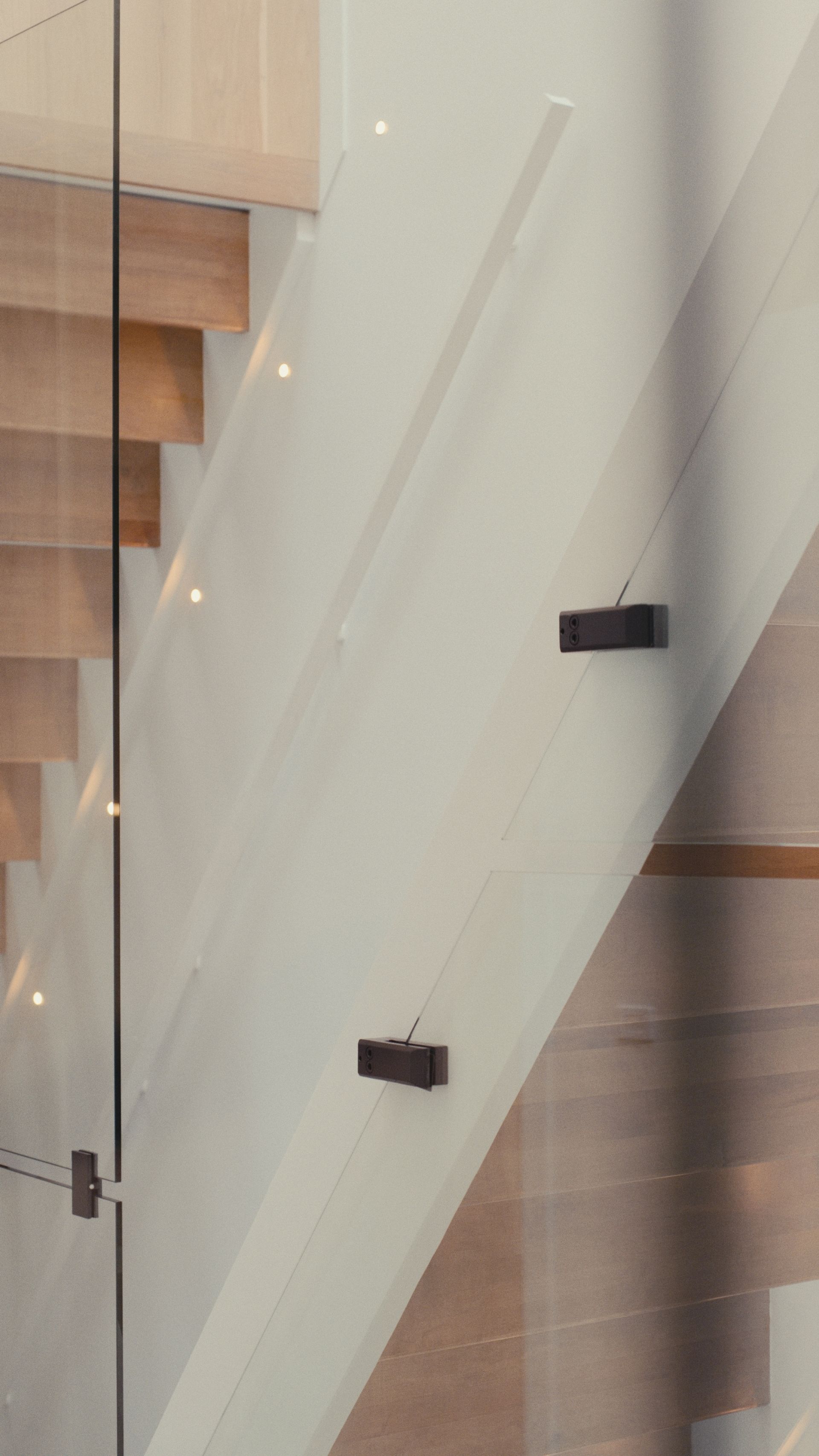 Escalier avec marches en bois et rambarde en verre fixé à un mur blanc avec des ferrures rectangulaires foncées.