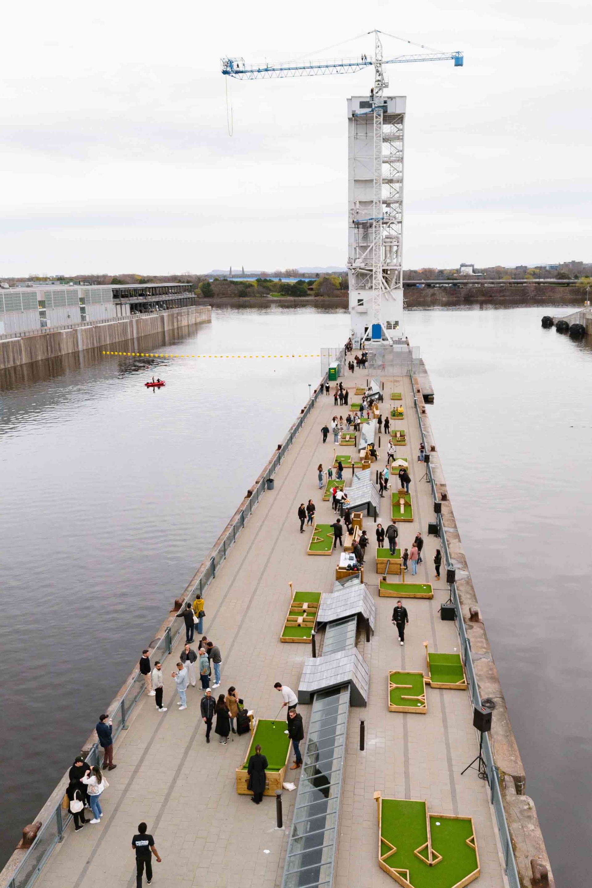 Des personnes marchent sur un quai en direction d'une haute grue à tour surplombant une eau calme.