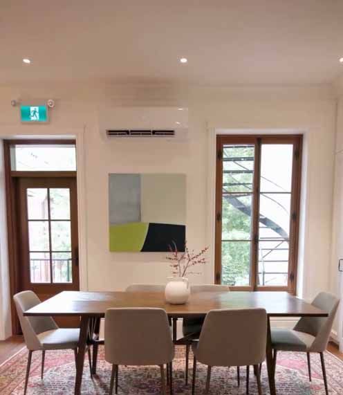Salle à manger moderne avec table en bois, chaises rembourrées, décorations murales et grandes fenêtres laissant entrer la lumière naturelle.