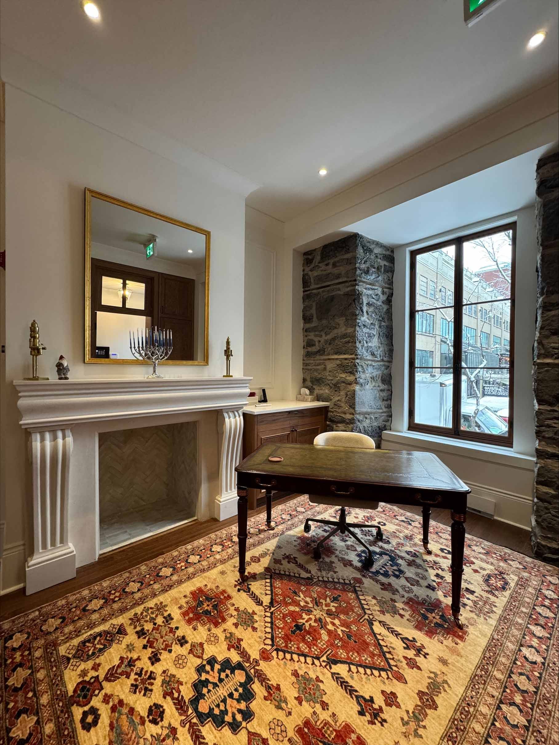 Élégant salon avec cheminée blanche, miroir, bureau en bois, tapis à motifs et coin salon sous la fenêtre en baie.