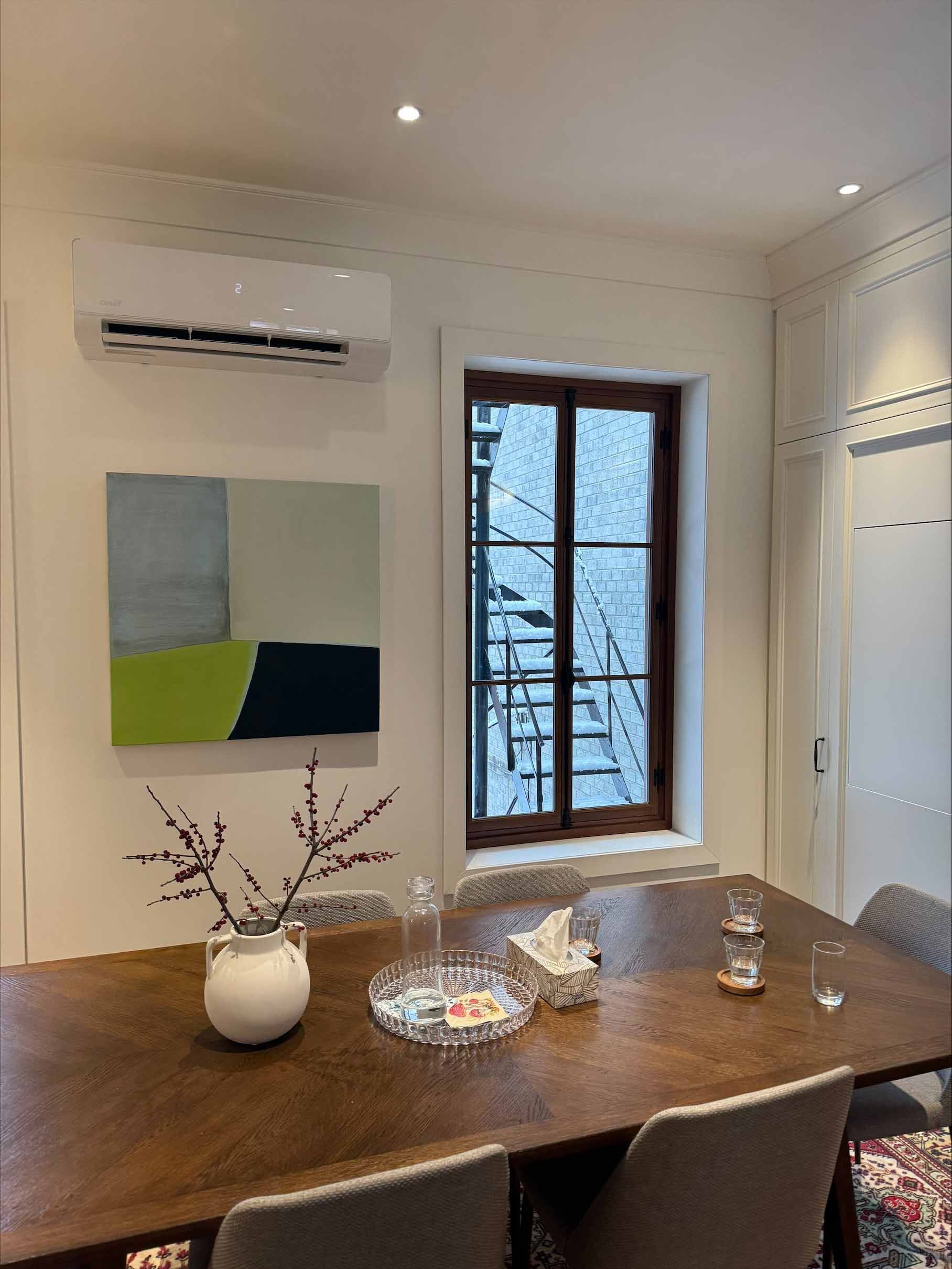 Salle à manger moderne avec table en bois, art mural abstrait et fenêtre donnant sur des arbres enneigés.