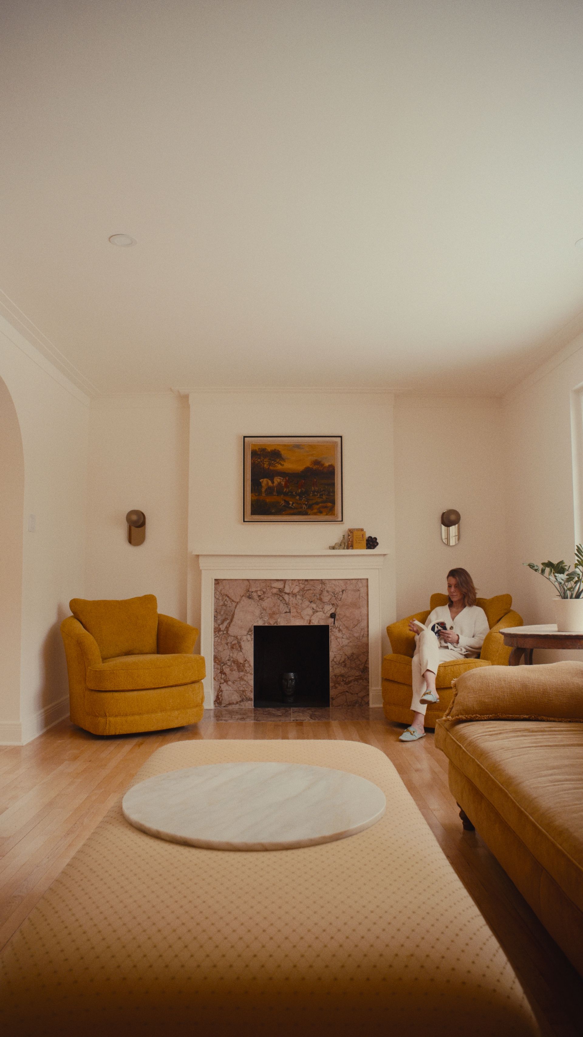 Une personne est assise dans un fauteuil jaune moutarde, dans un salon avec une cheminée et un parquet.