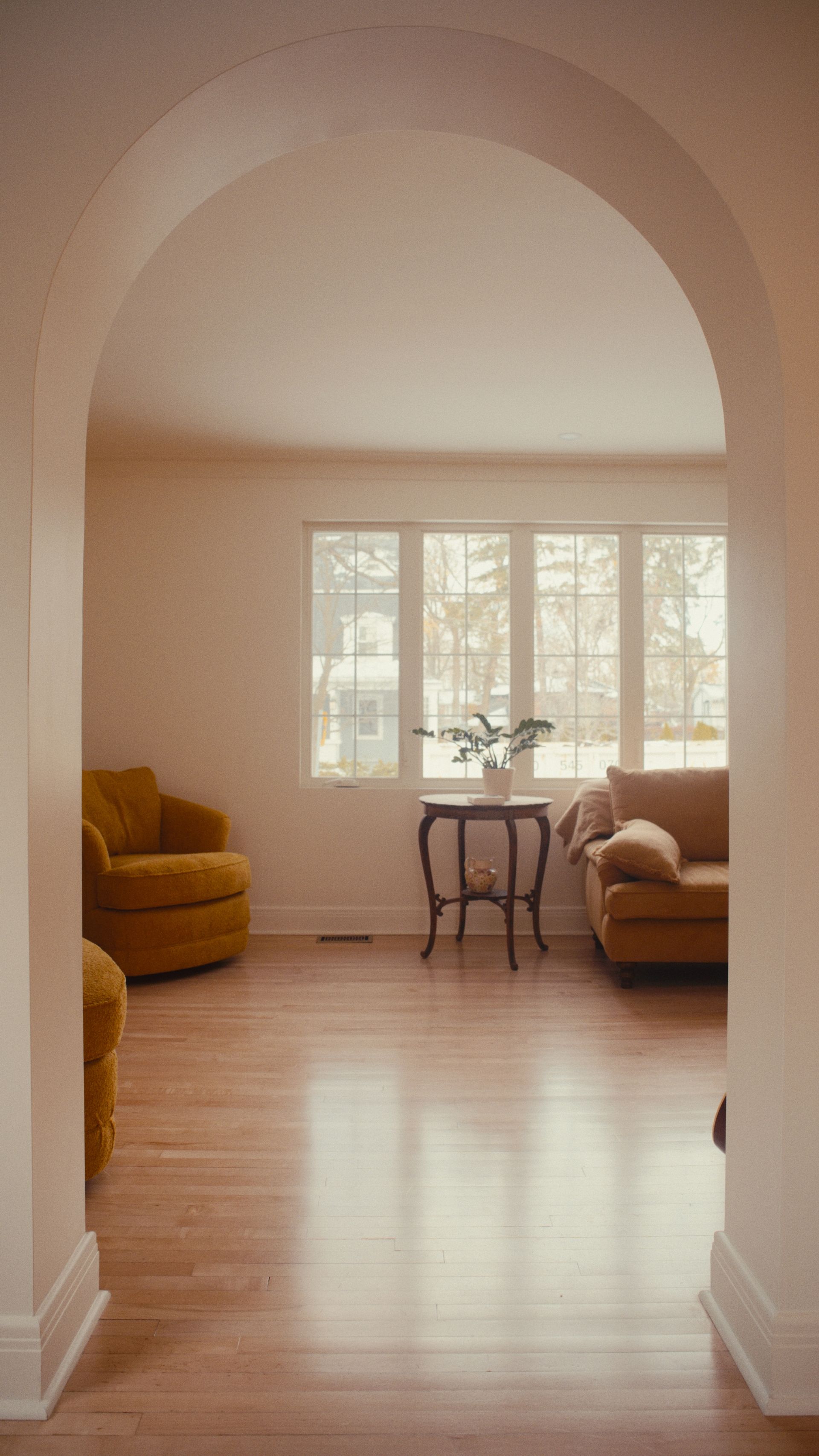 Une arche donne accès à un salon avec parquet, meubles couleur moutarde et une fenêtre laissant entrer la lumière naturelle.
