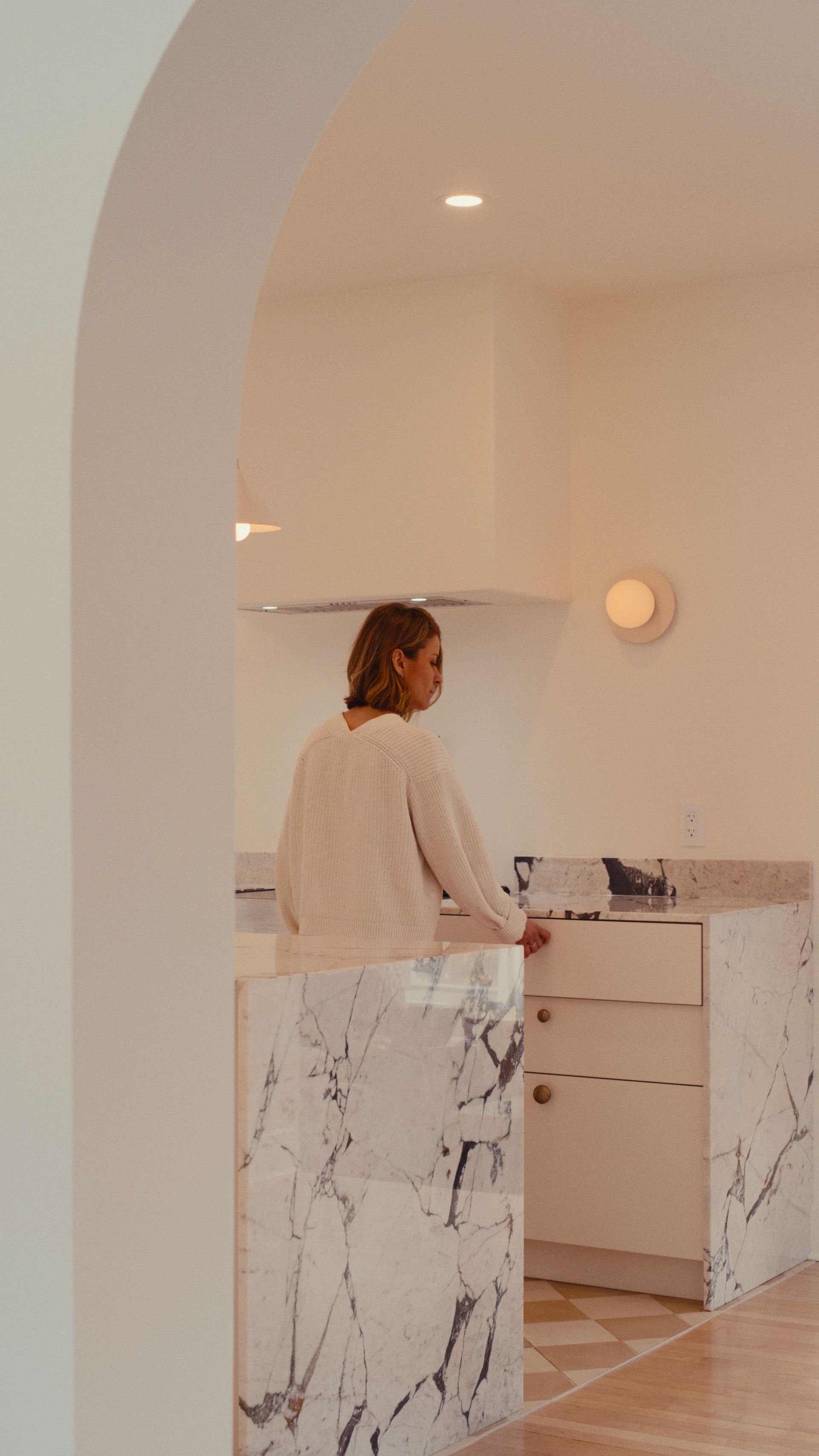 Une femme ouvre un tiroir dans une cuisine moderne avec un îlot en marbre et une porte cintrée.
