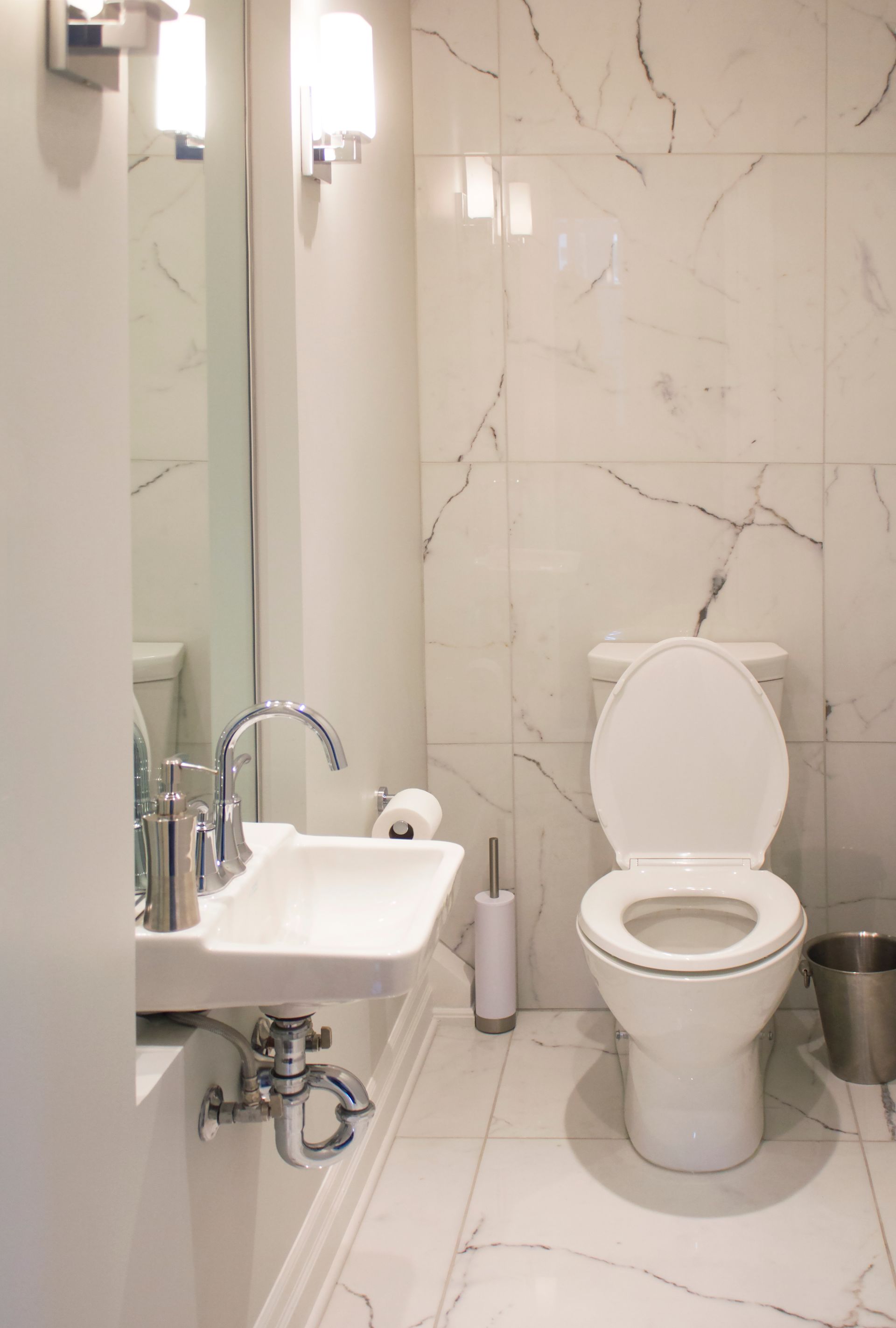 Salle de bains avec murs et sol carrelés façon marbre, toilettes blanches à abattant ouvert et petit lavabo.