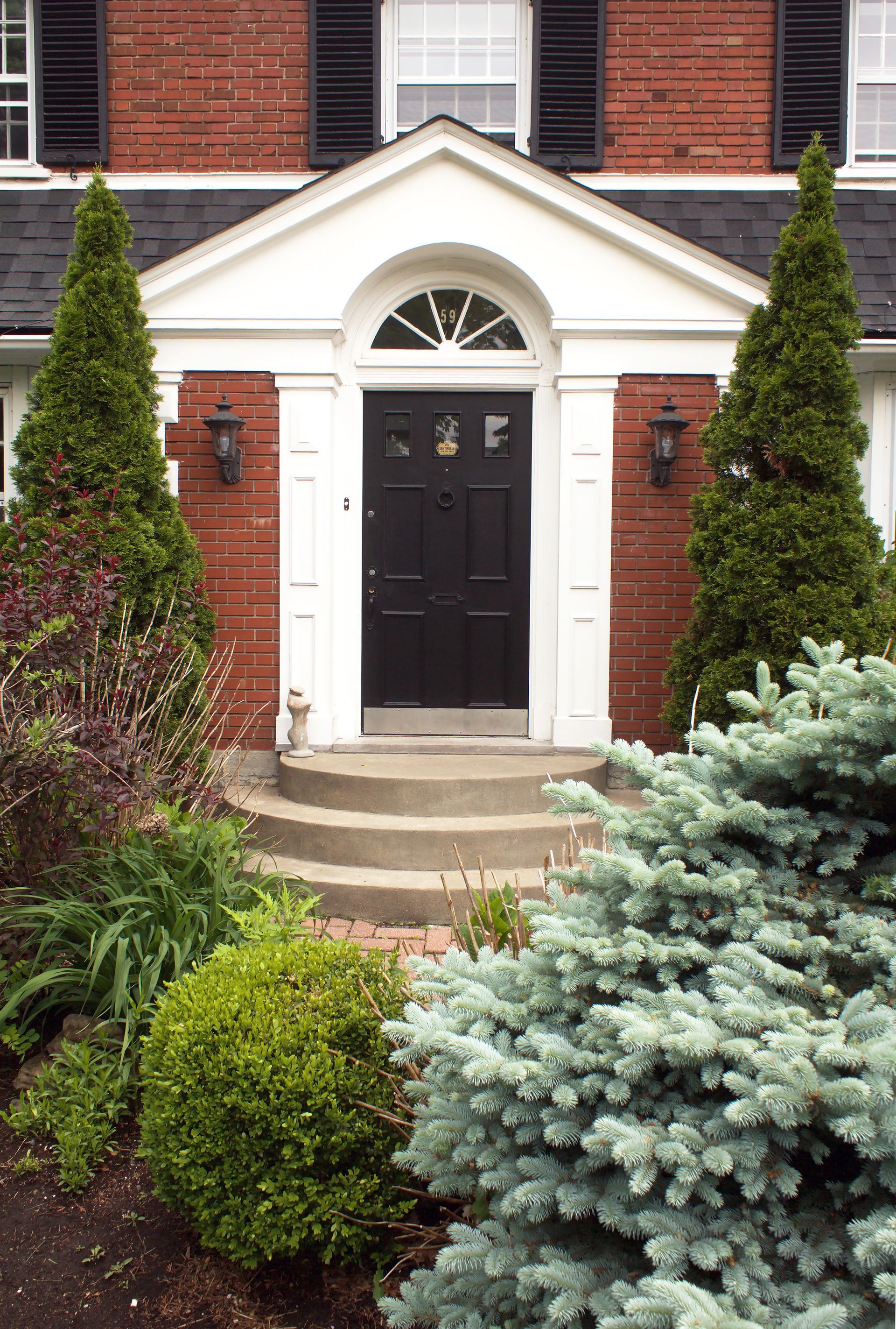 Entrée d'une maison en briques avec porte noire, encadrements blancs et aménagement paysager avec arbustes et arbres.