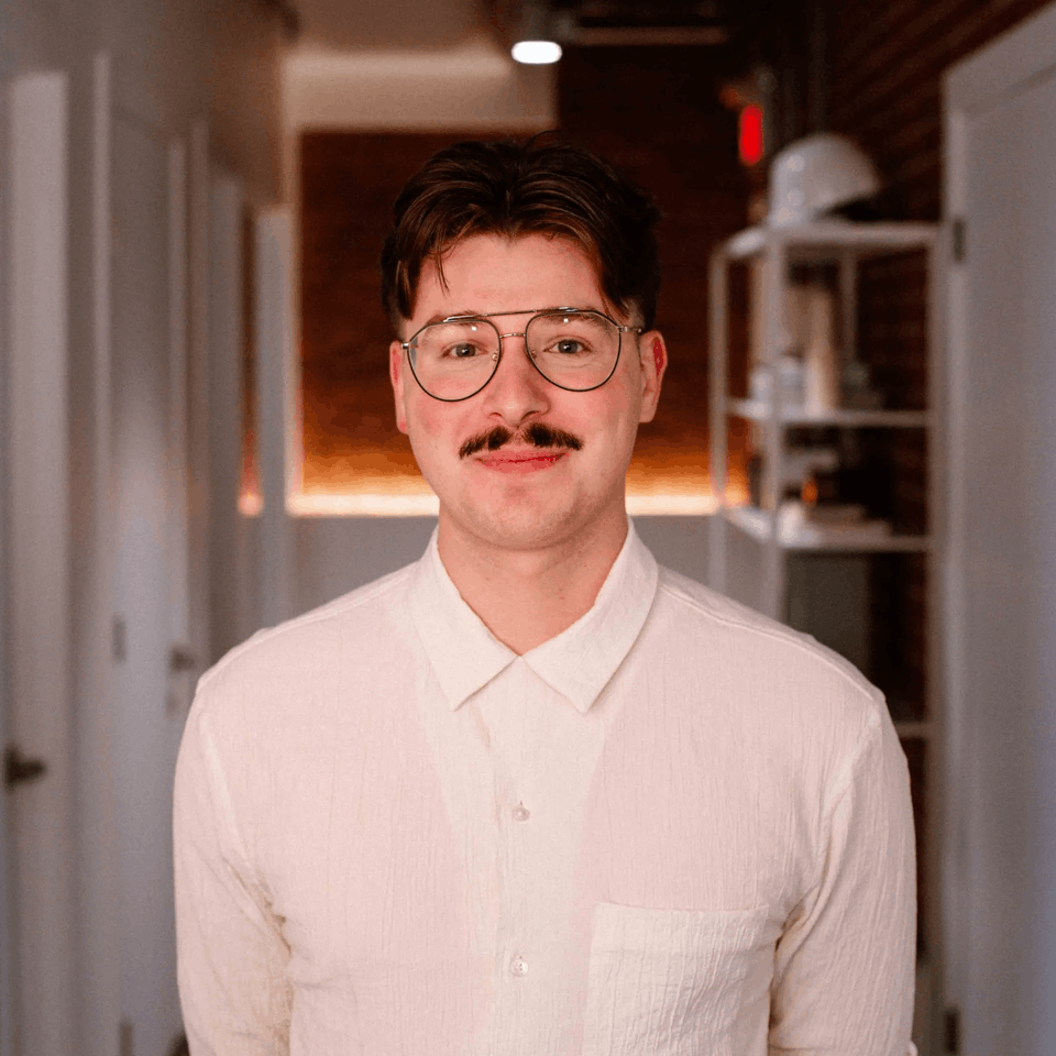 Portrait d'un homme portant des lunettes et une moustache, vêtu d'une chemise blanche, dans un couloir faiblement éclairé.
