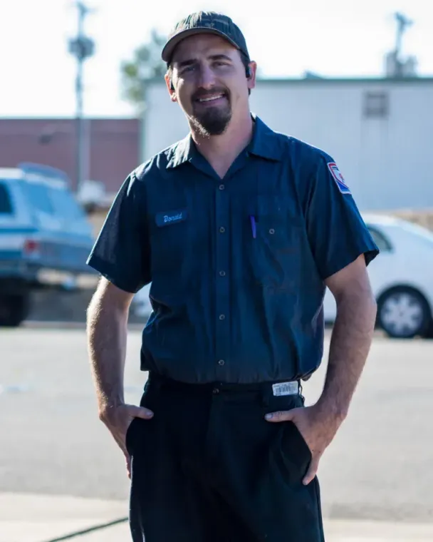 Mechanic in dark blue uniform and cap smiling outdoors, standing in a parking lot.