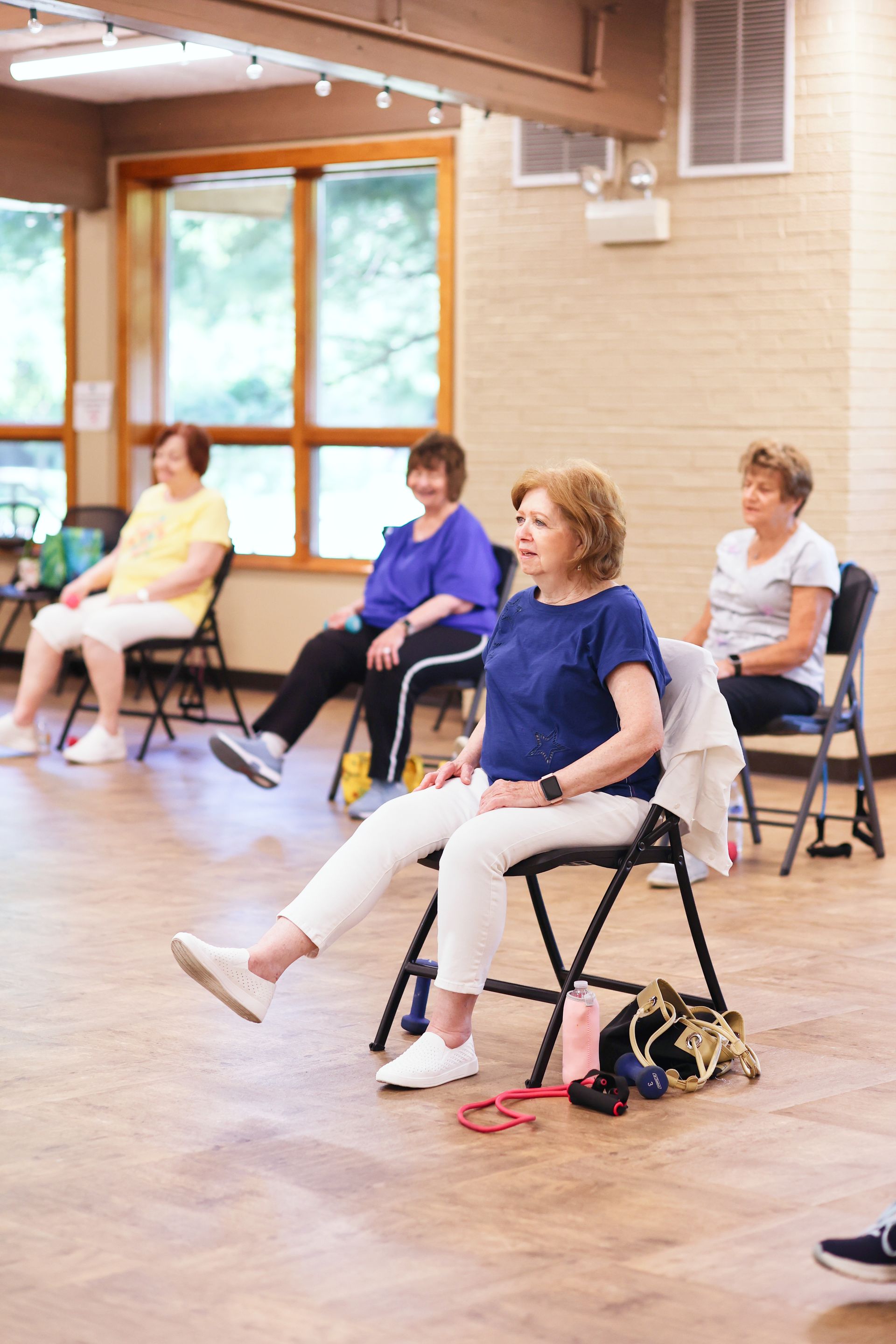 People seated on folding chairs in a bright studio, exercising with raised legs during a class.