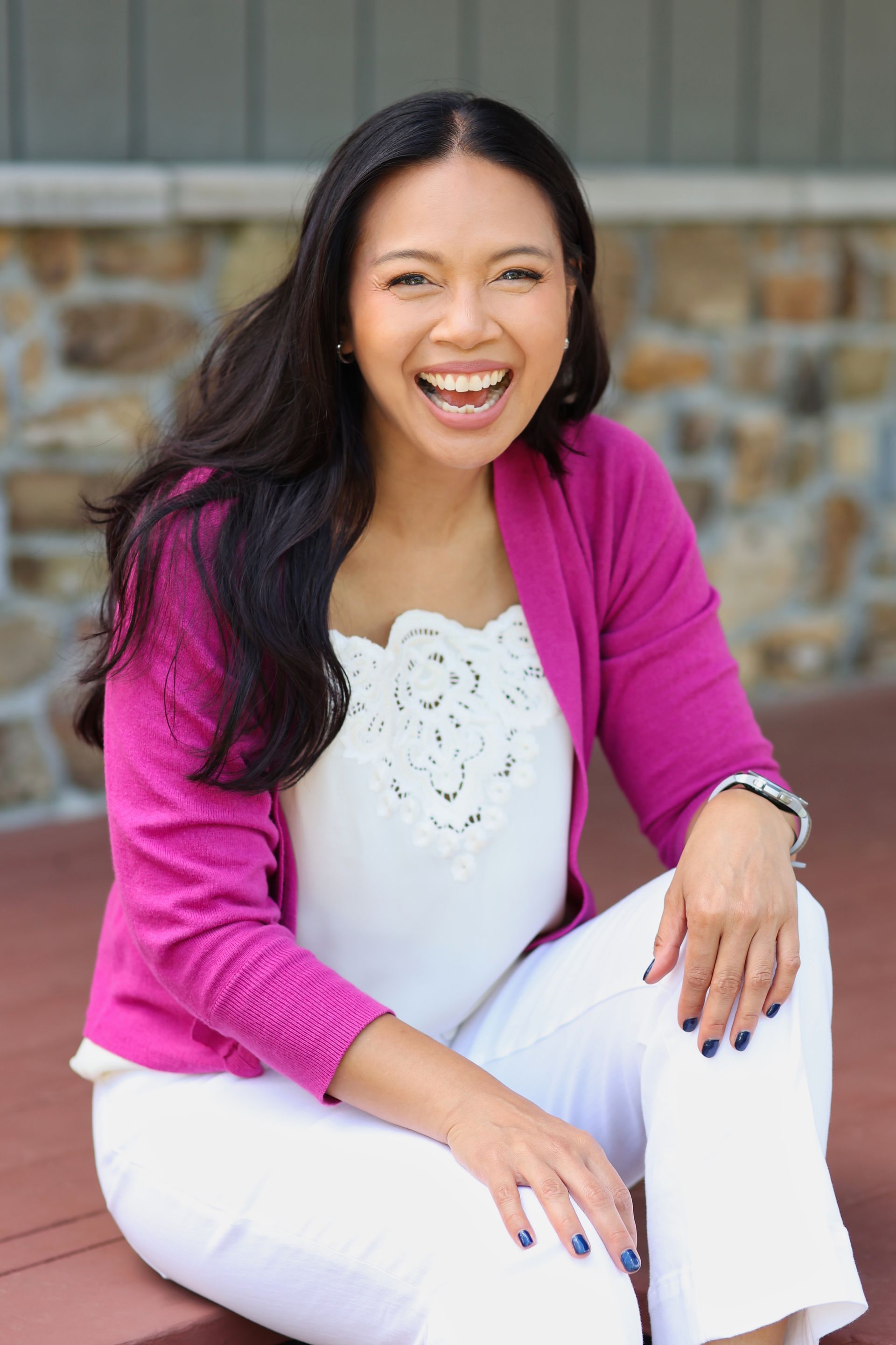 Smiling woman in a magenta cardigan and white outfit sitting outdoors against a stone wall