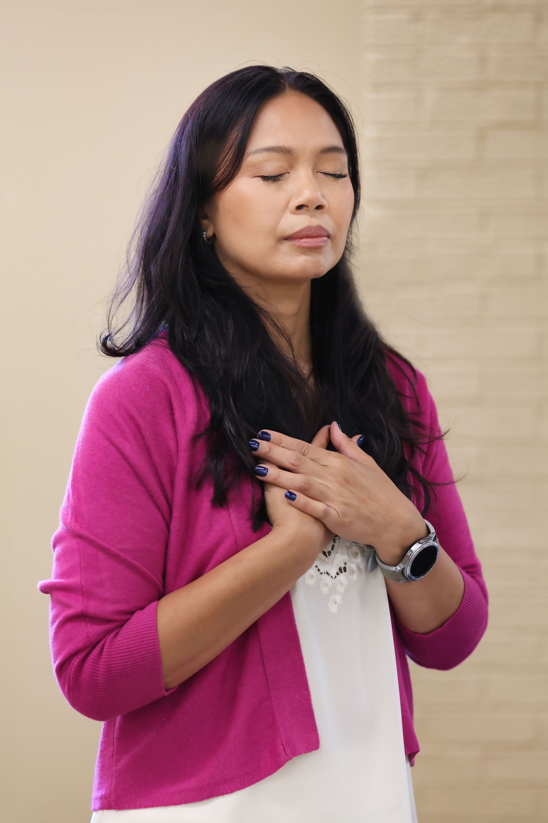 Woman in a pink cardigan with eyes closed, hands over chest, standing indoors against a beige brick wall.