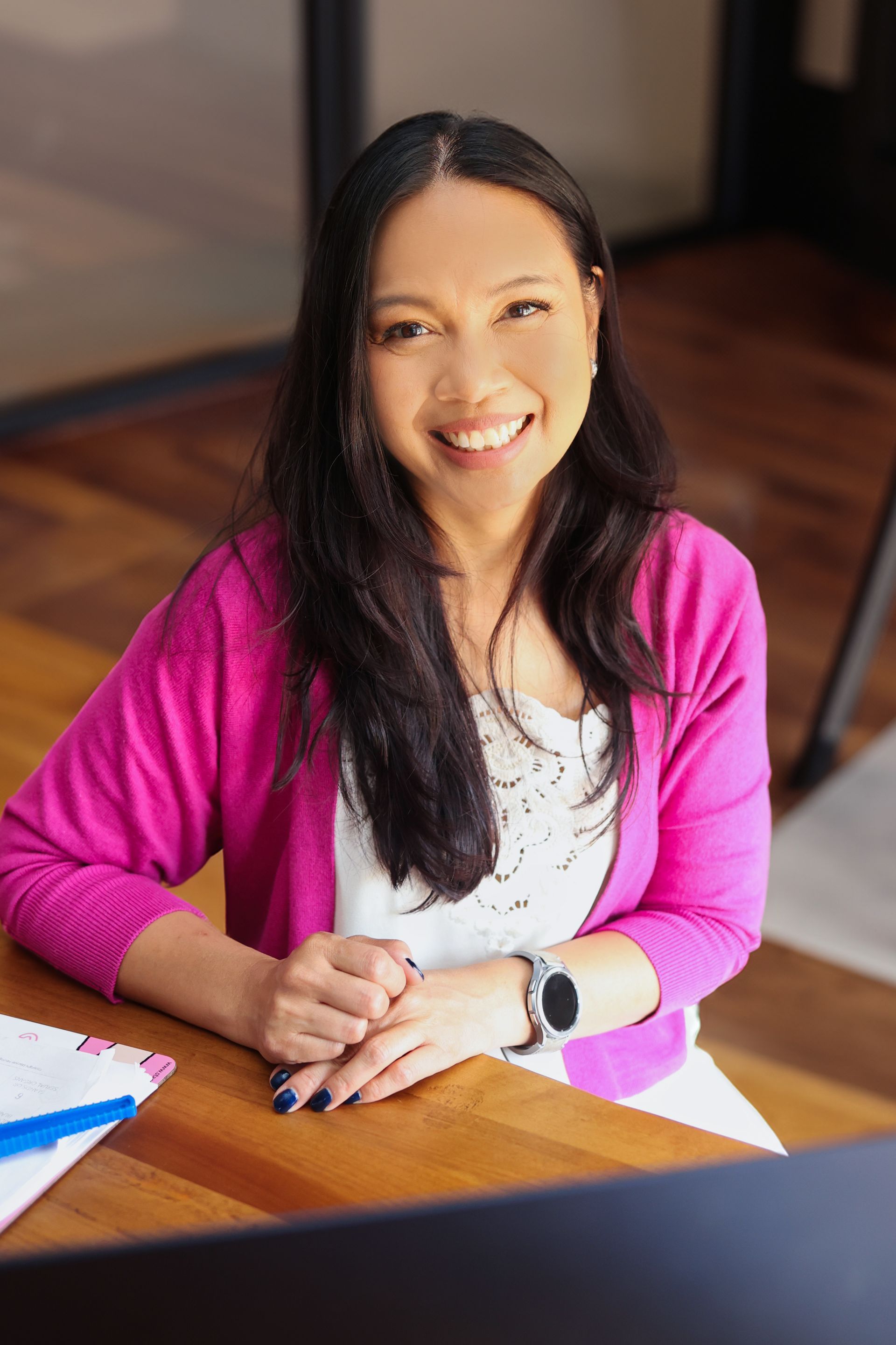 Smiling woman in a pink cardigan seated at a wooden table in a bright office setting