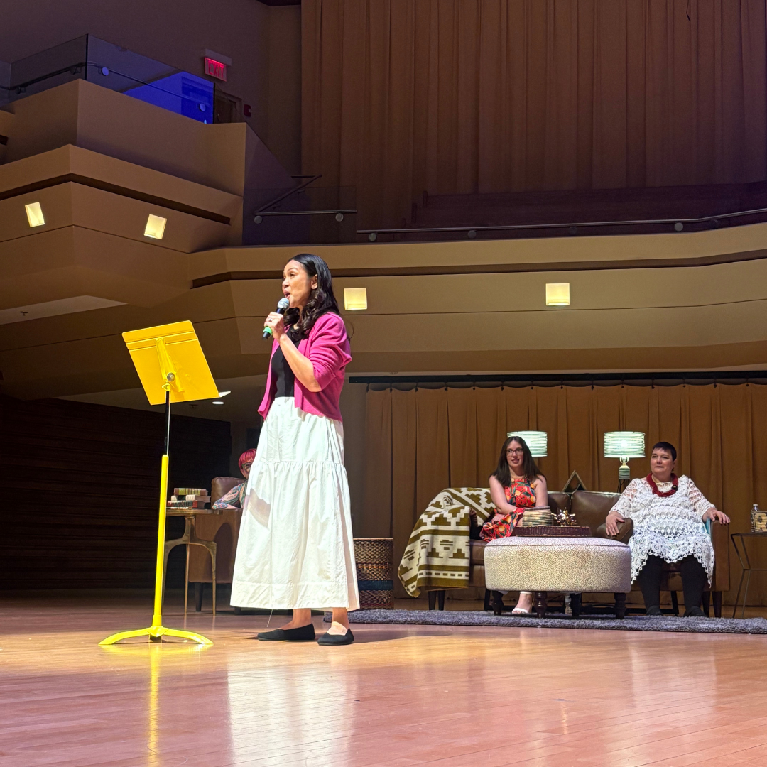 Woman singing on a concert hall stage beside a yellow music stand, with seated people in the background.