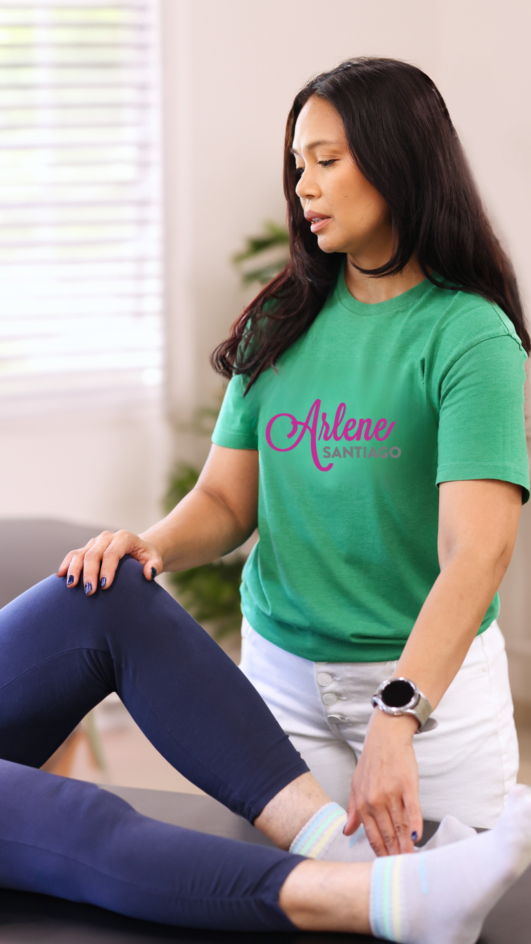 Woman in green T-shirt stretching another person’s leg in a bright therapy room