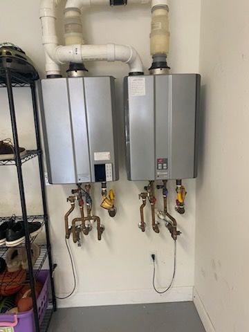 Two wall-mounted, silver tankless water heaters with copper pipes and white vent pipes in a garage.