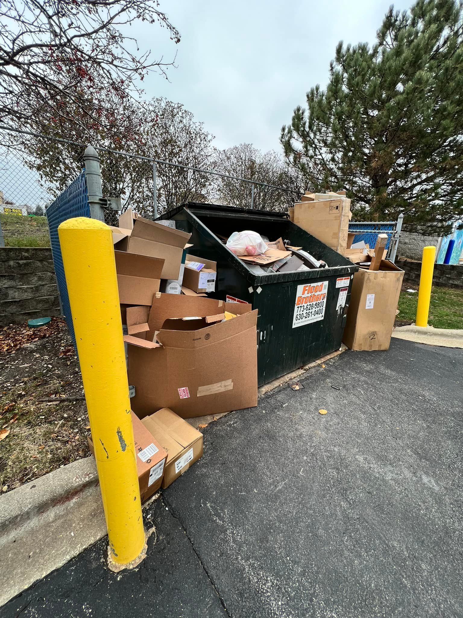 Dumpster overflowing with trash and cardboard boxes in an outdoor area, next to yellow bollards. Overcast sky in the background.