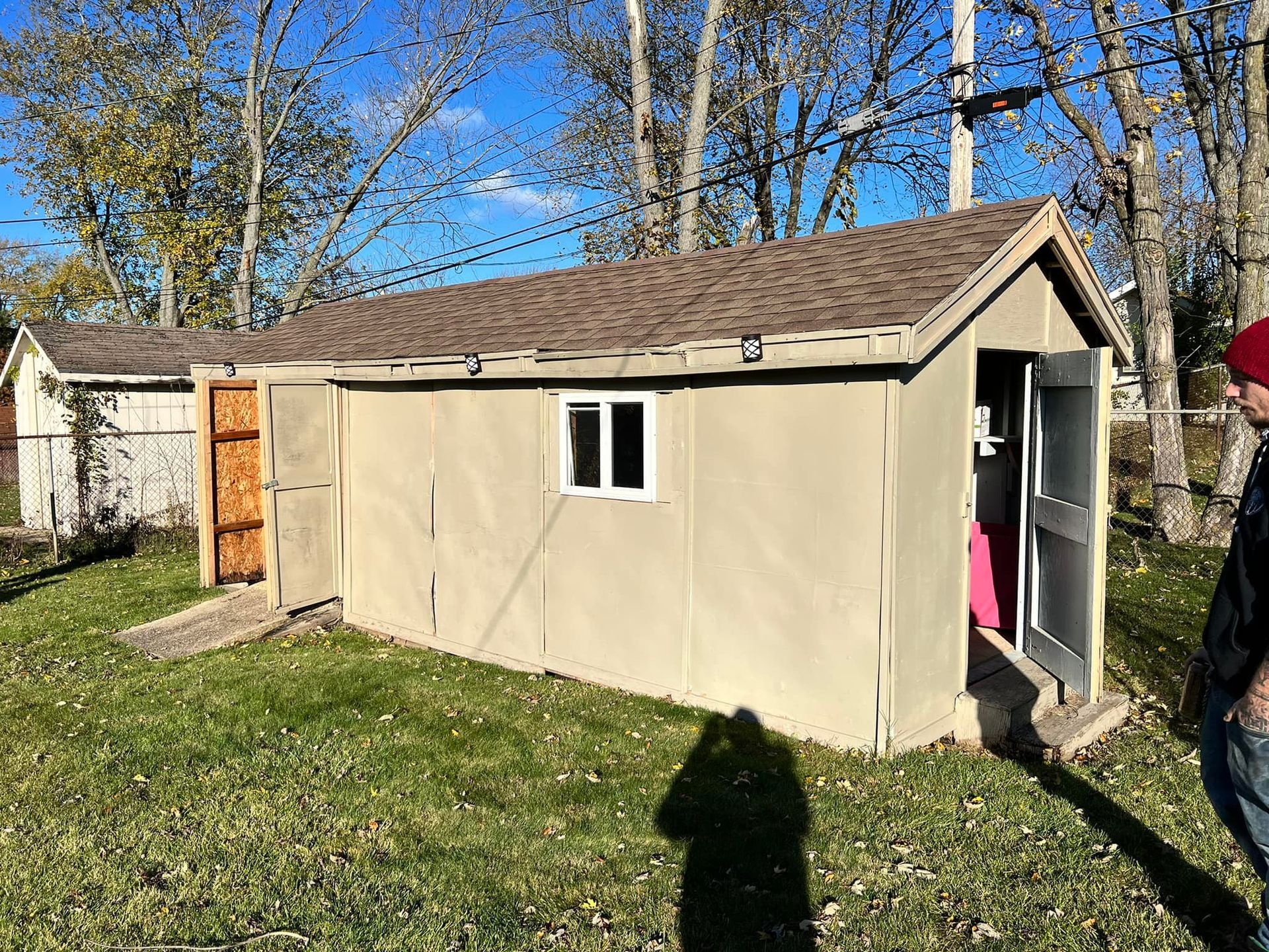Tan shed with brown roof, white-framed window, and open doors, standing on green grass. A person stands to the right.