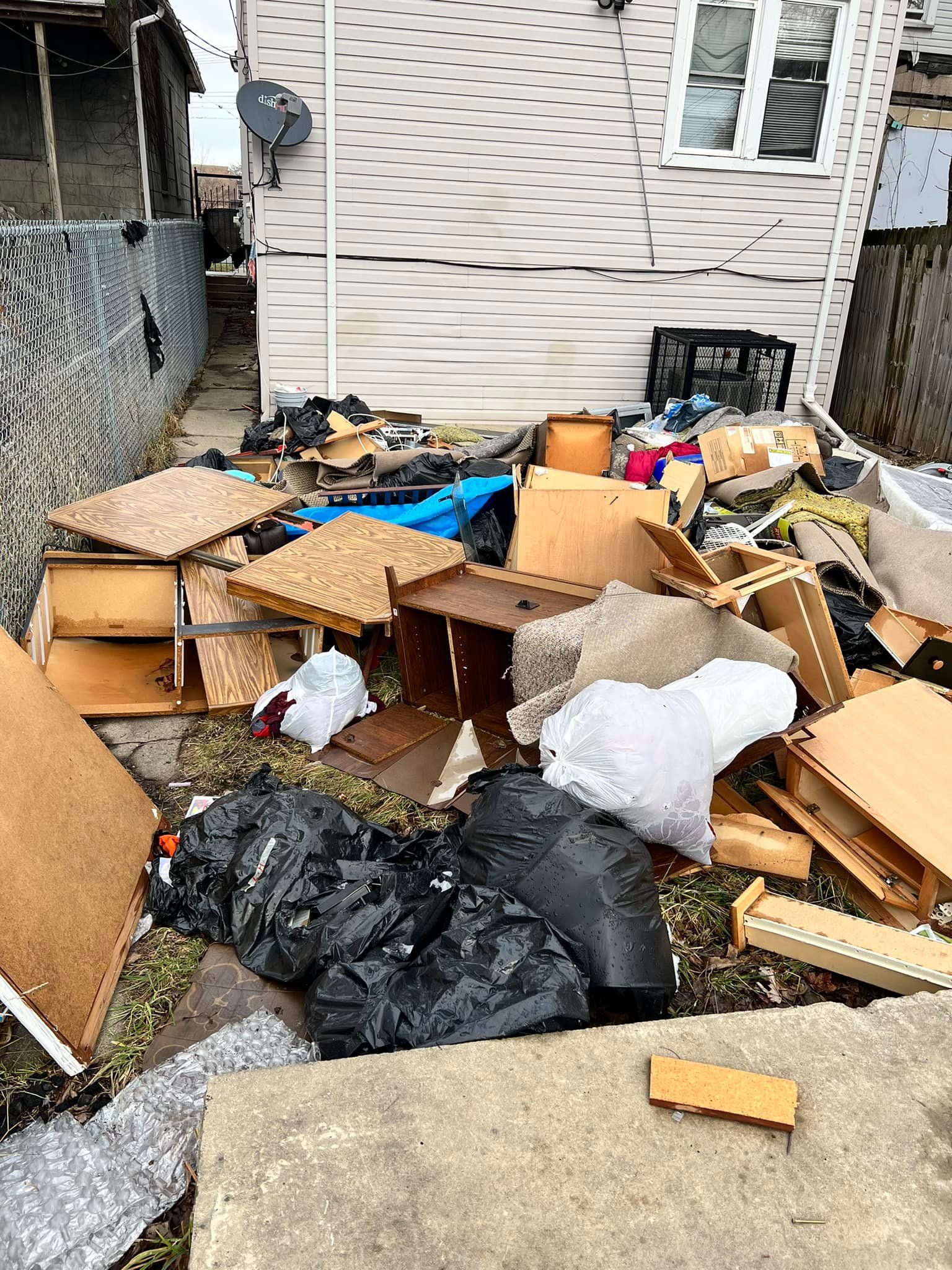Backyard with a large pile of discarded trash, including furniture, boxes, and bags, near a weathered house.