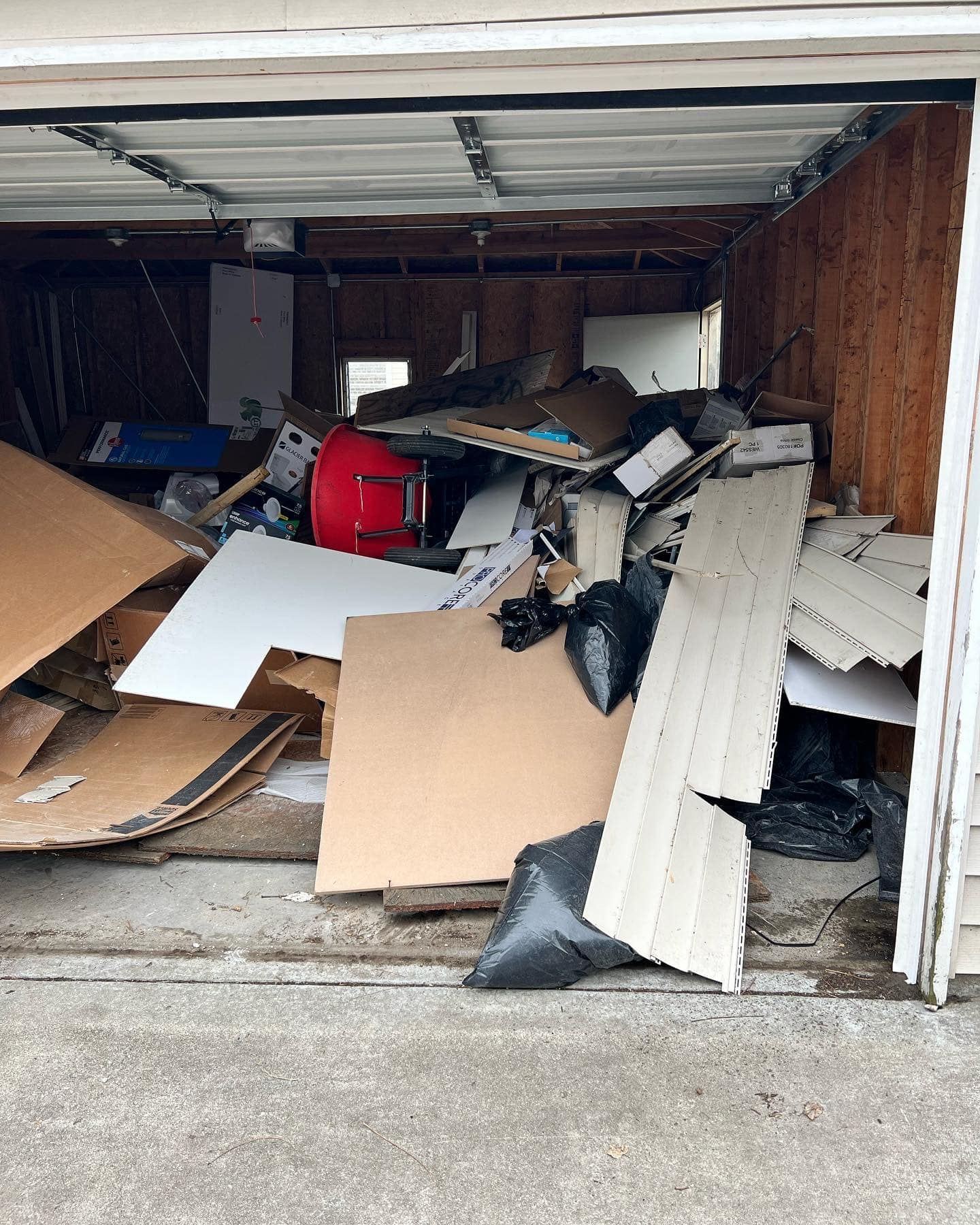 A garage filled with a large pile of debris, including cardboard, wood, and trash bags. A red drum is visible.