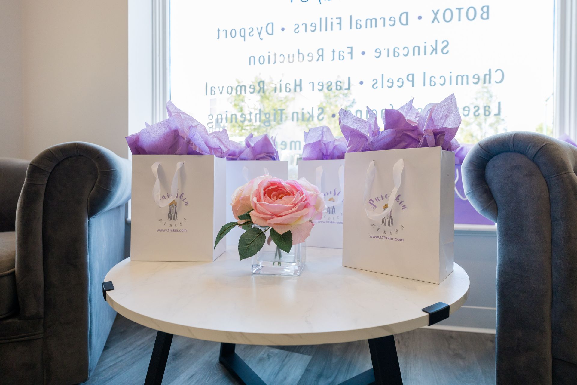 Gift bags and a pink flower arrangement on a round table by a window, framed by two armchairs