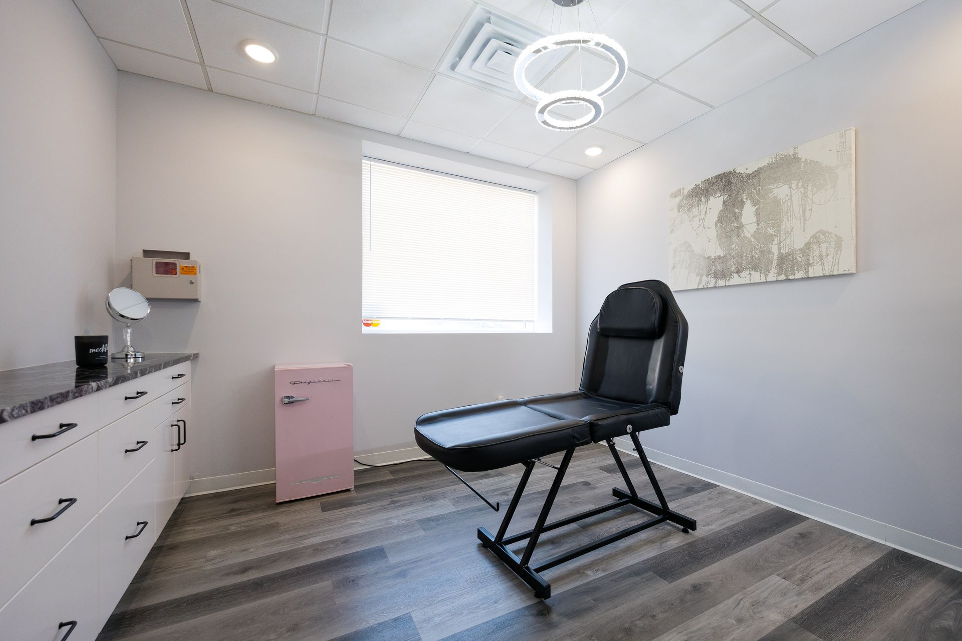 Treatment room with black exam chair, white cabinets, window blinds, and soft pink storage cabinet