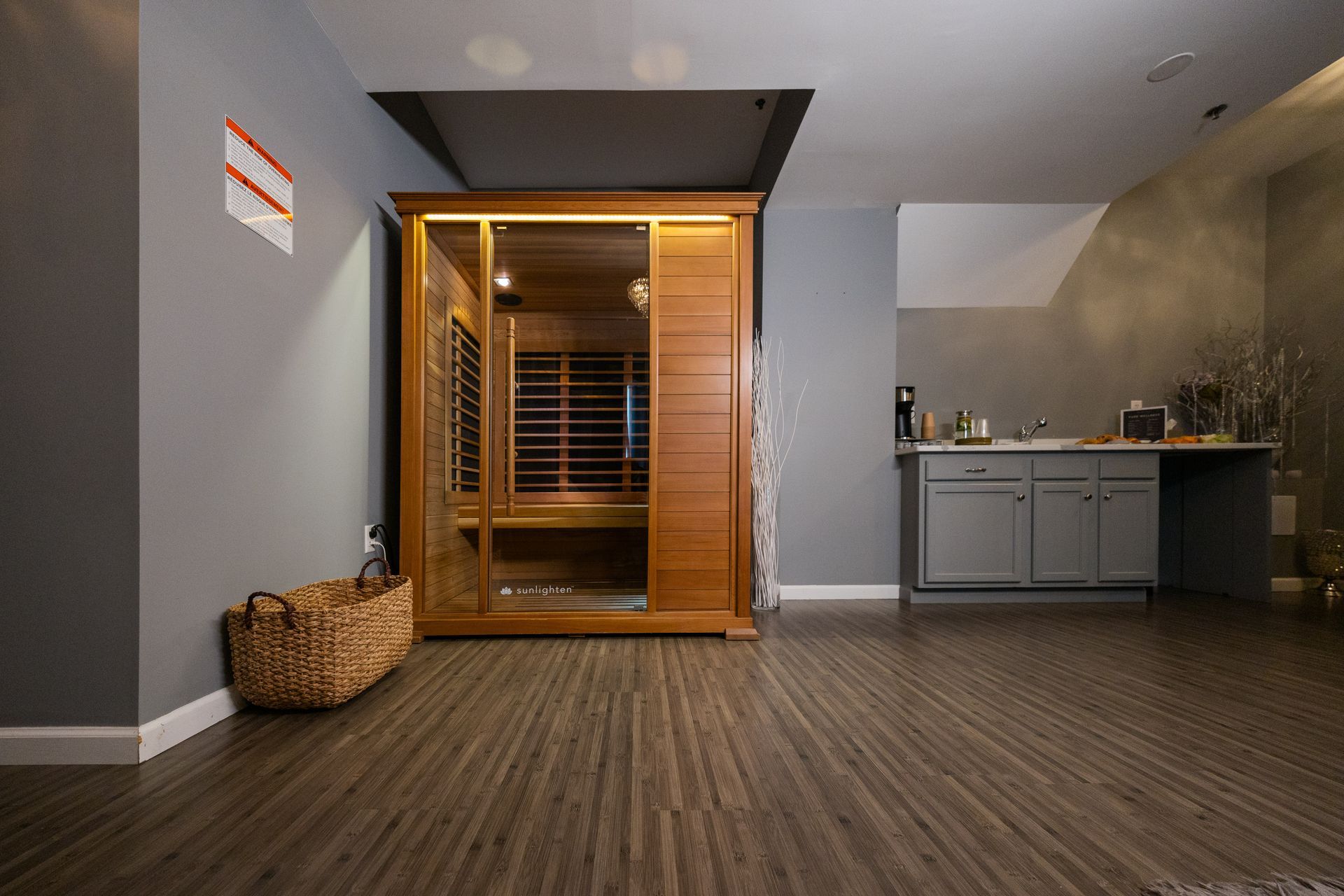 Empty room with wooden sauna, wicker basket, and gray kitchenette on dark wood flooring