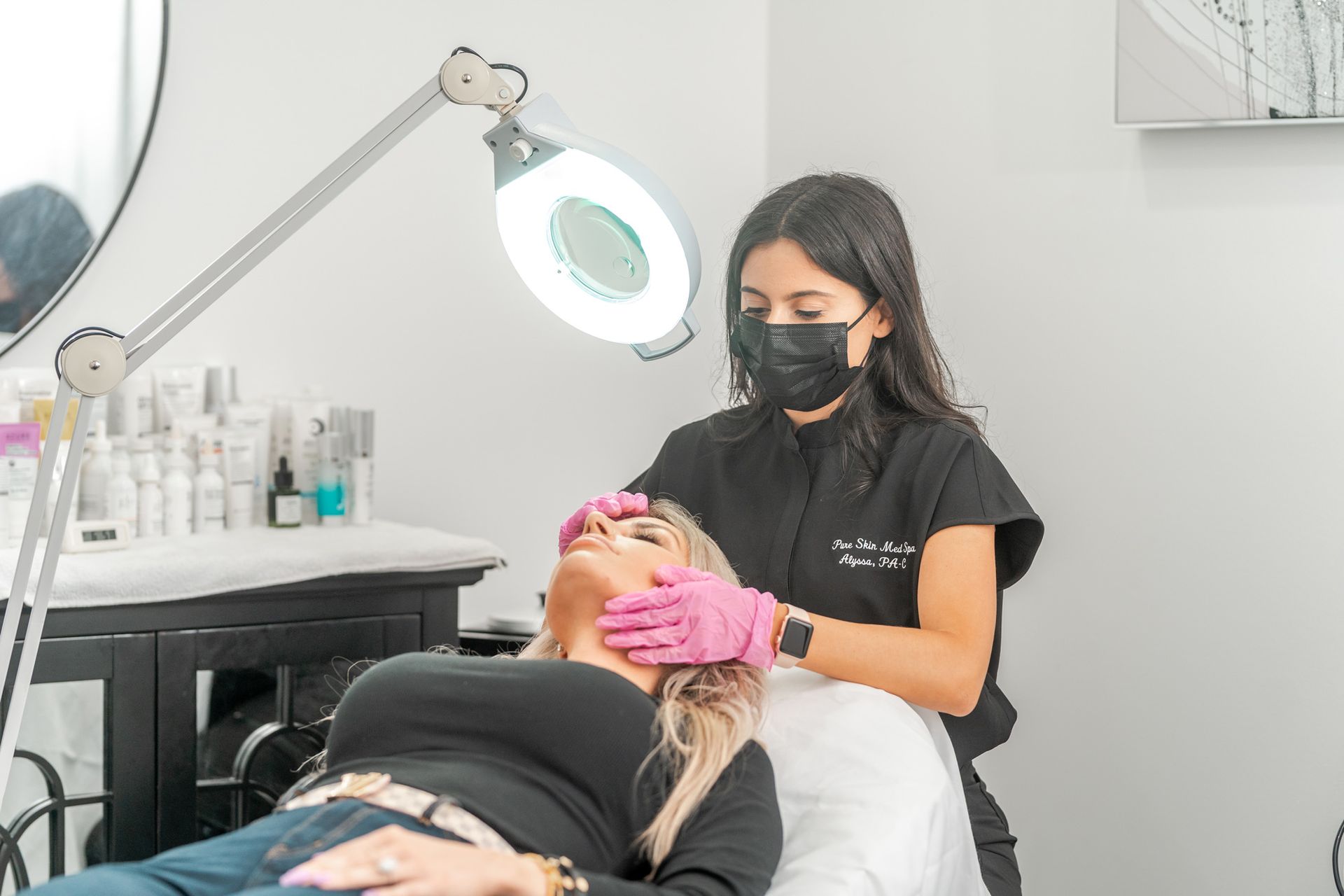 Technician in black mask applies eyelash treatment to client on a salon bed under a magnifying lamp