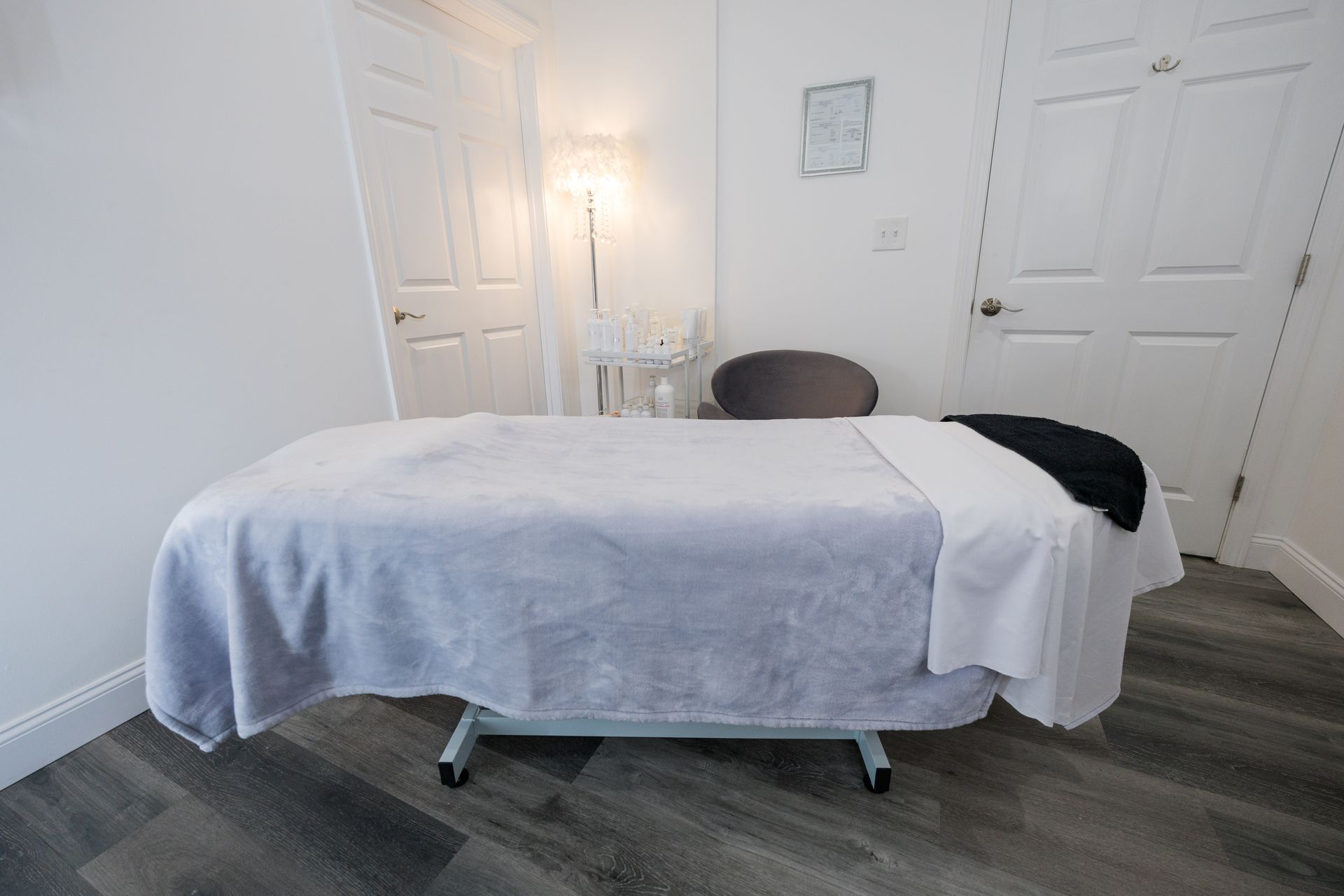 Massage table in a bright room with white doors, a lamp, chair, and gray wood floor.