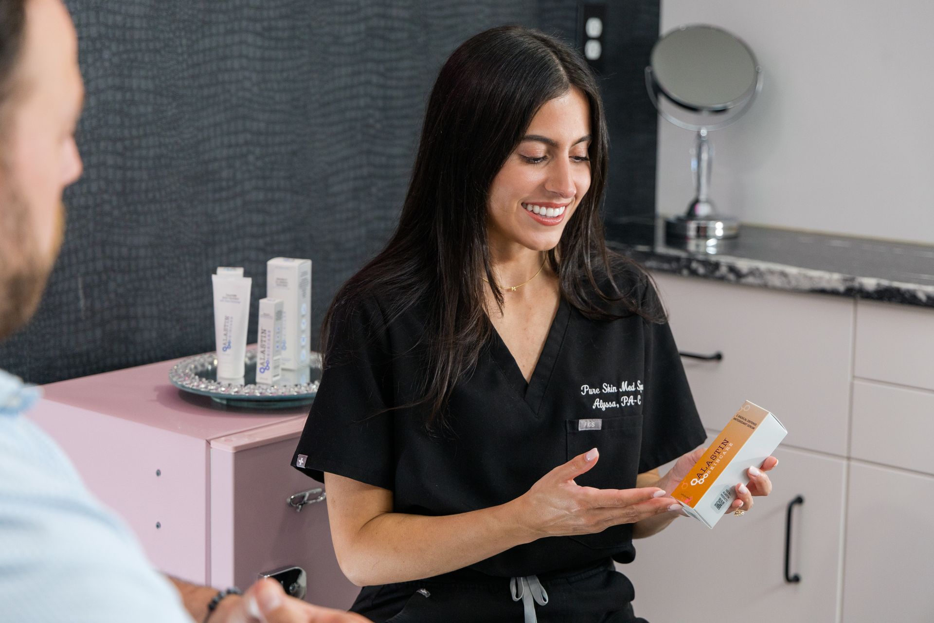 Woman in black scrubs smiling while holding a product in a clinic room