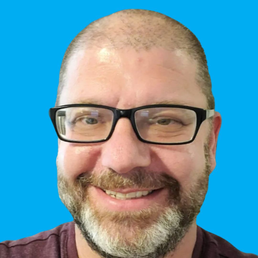 Bald man with glasses and a beard smiles at the camera against a blue background.