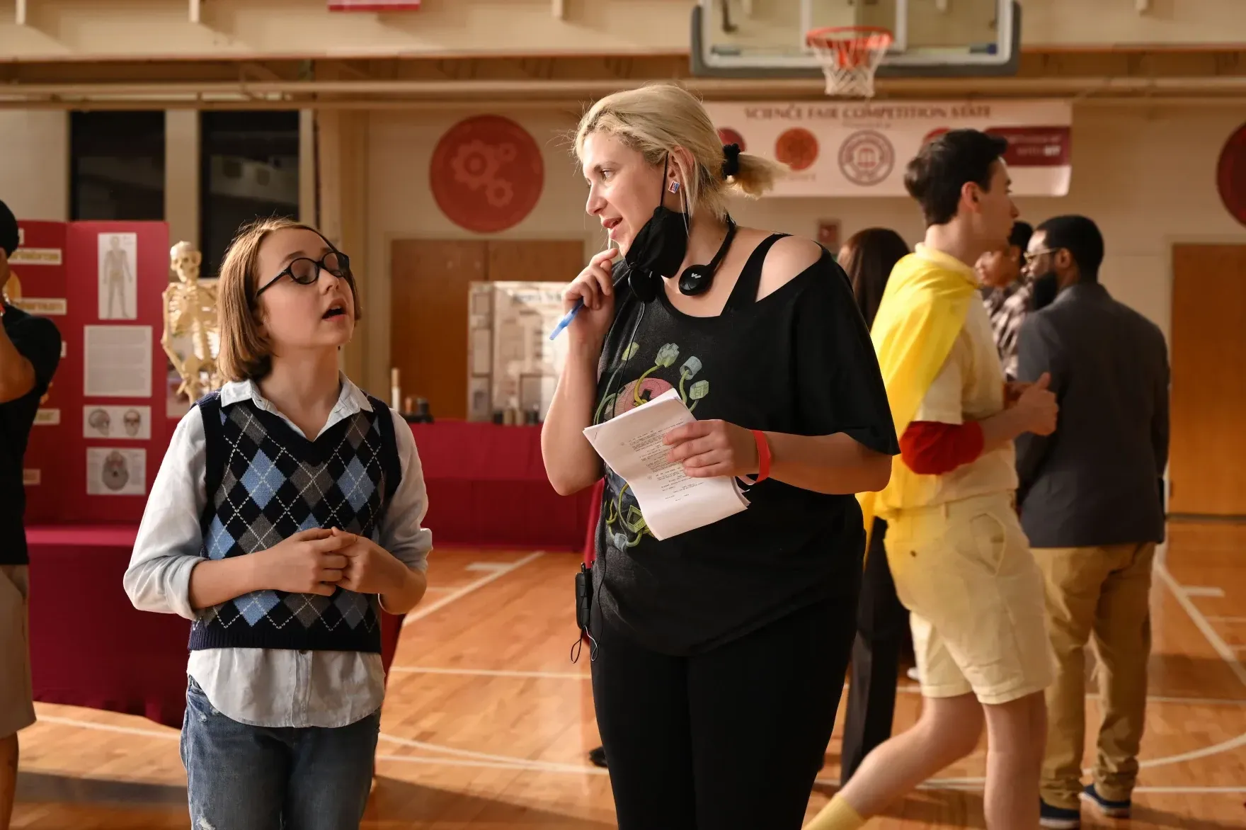 A woman with a microphone and a young boy wearing glasses stand in a gymnasium. Others in the background.