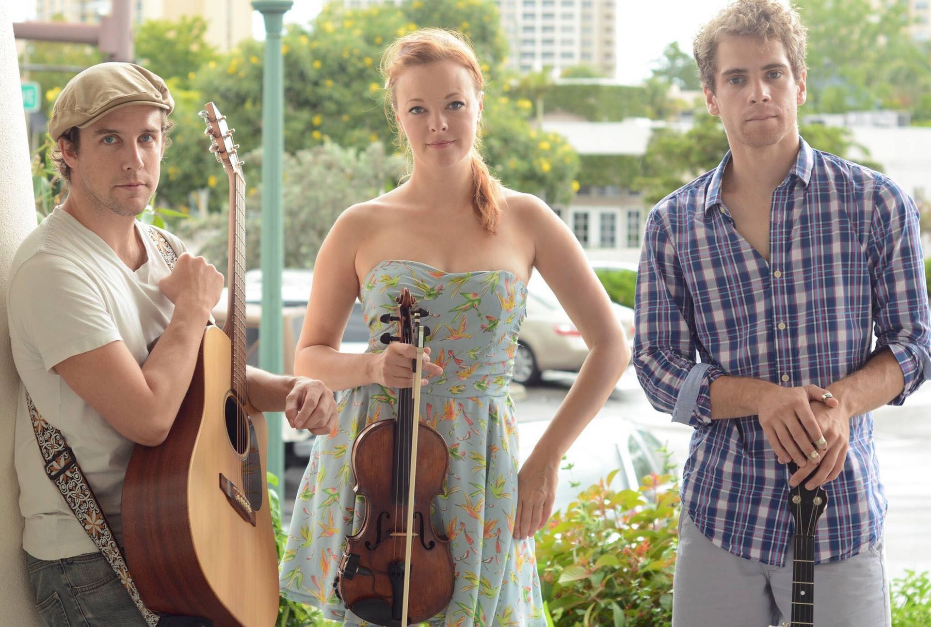 Three musicians posing outdoors: a man with guitar, a woman with violin, and a man with a mandolin.