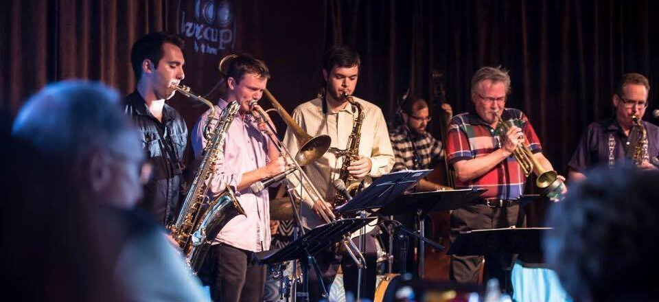 A jazz band performs on a stage, with several musicians playing brass and woodwind instruments in a dimly lit venue.