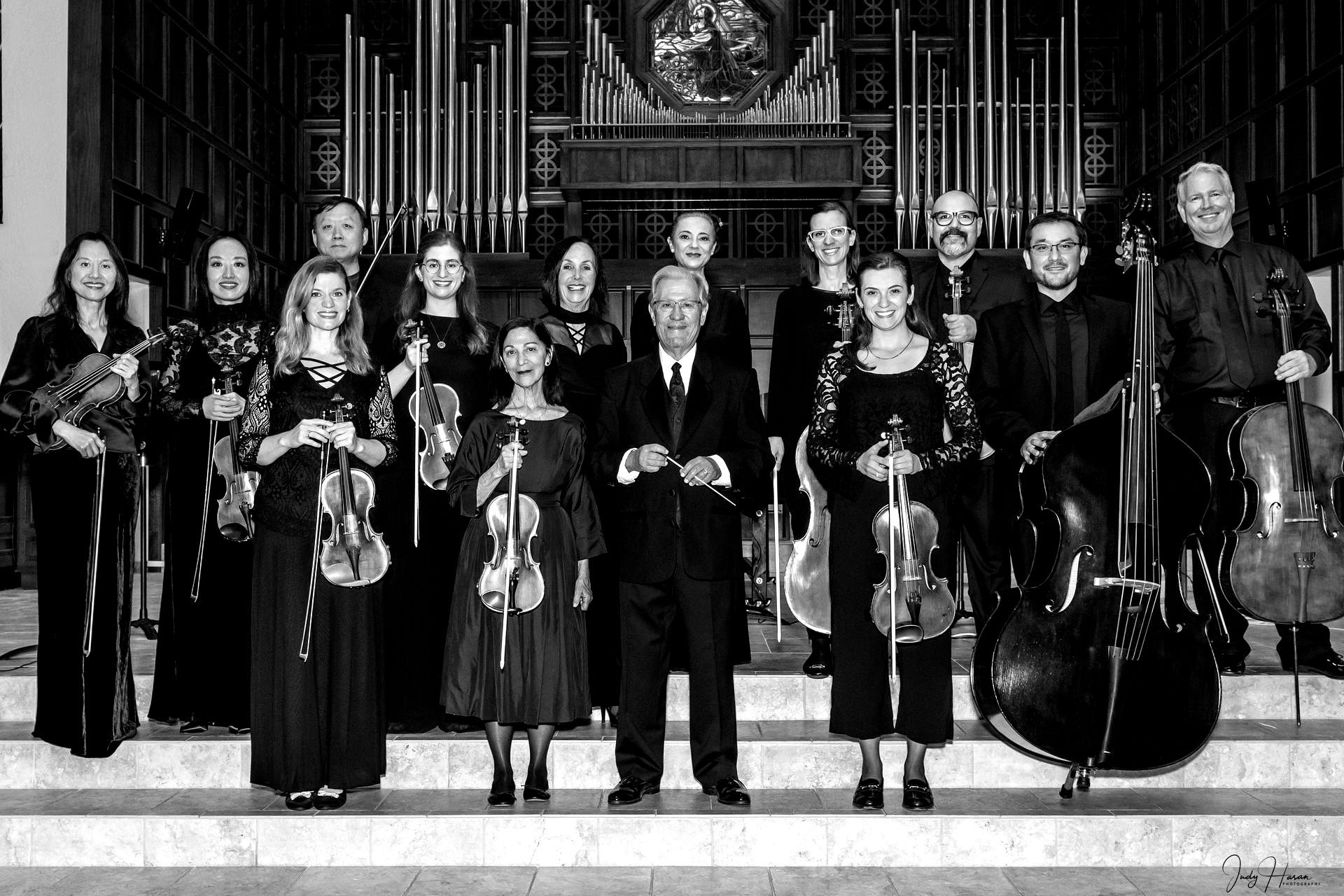 Orchestra in formal attire posing on steps in front of a church organ.