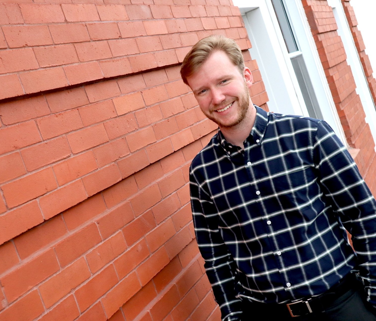 Man with red hair and beard, smiling in a blue and white plaid shirt, leans against a red brick wall.