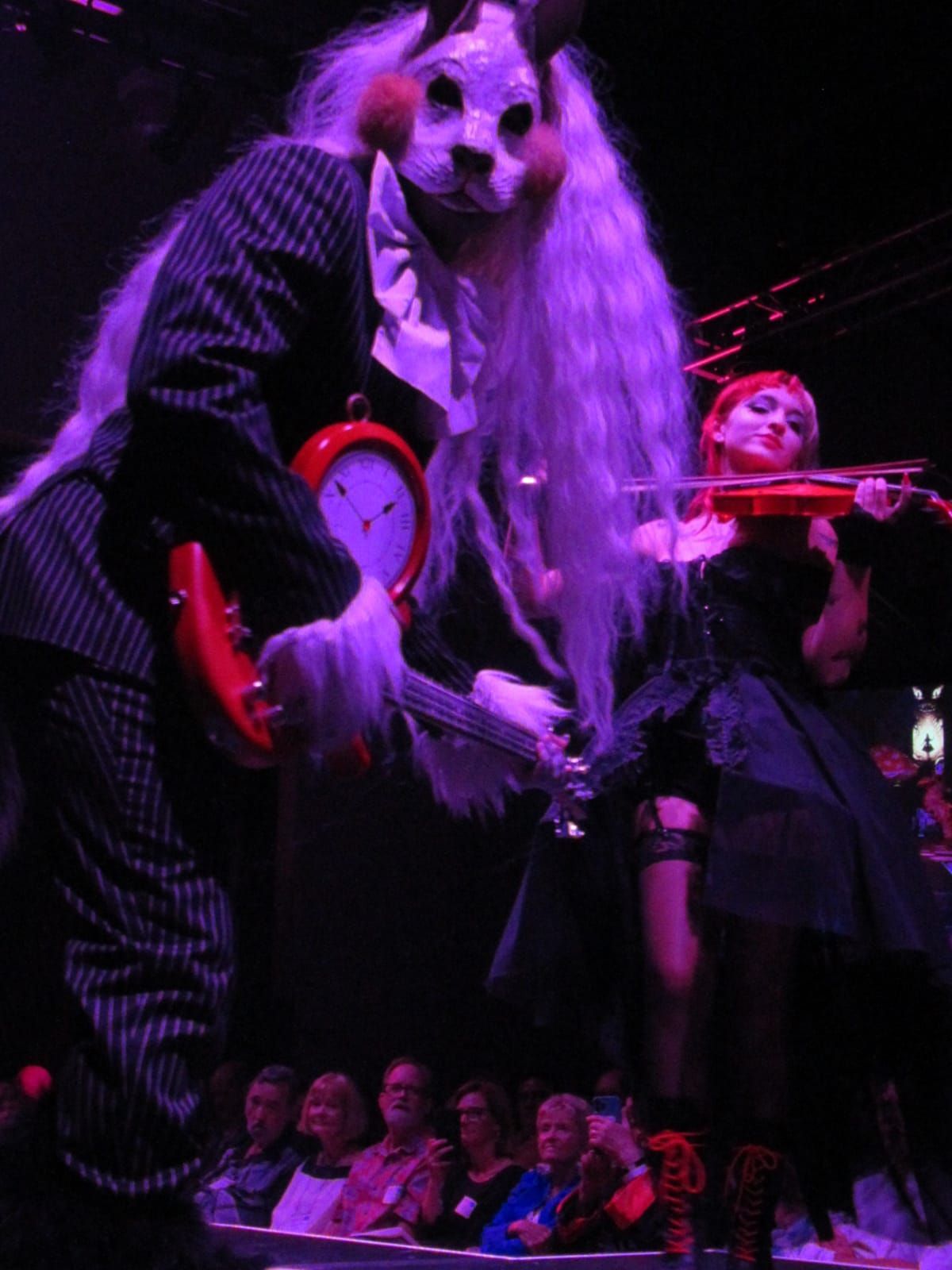 A giant rabbit in pinstripes holds a clock, next to a person playing violin. Dark stage, audience visible.