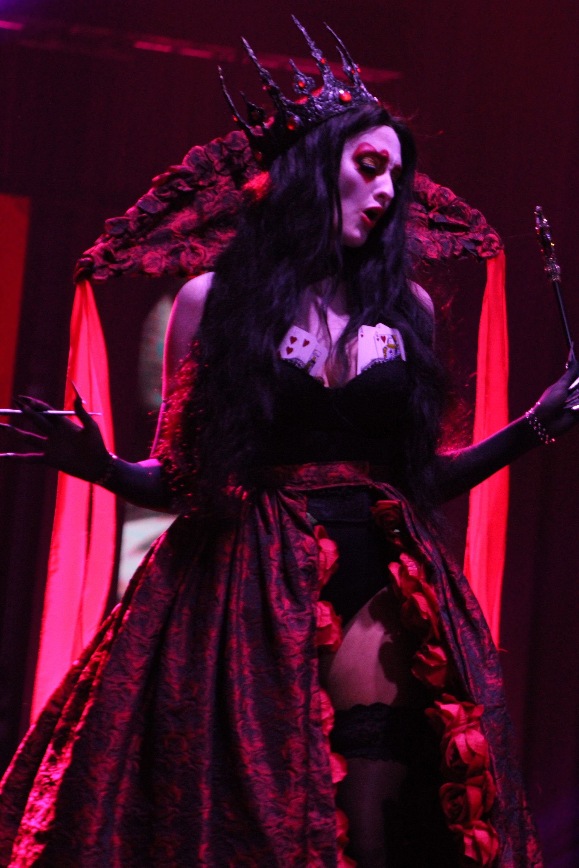 Woman in black gothic queen attire, wearing a crown, striking a pose, red lighting.