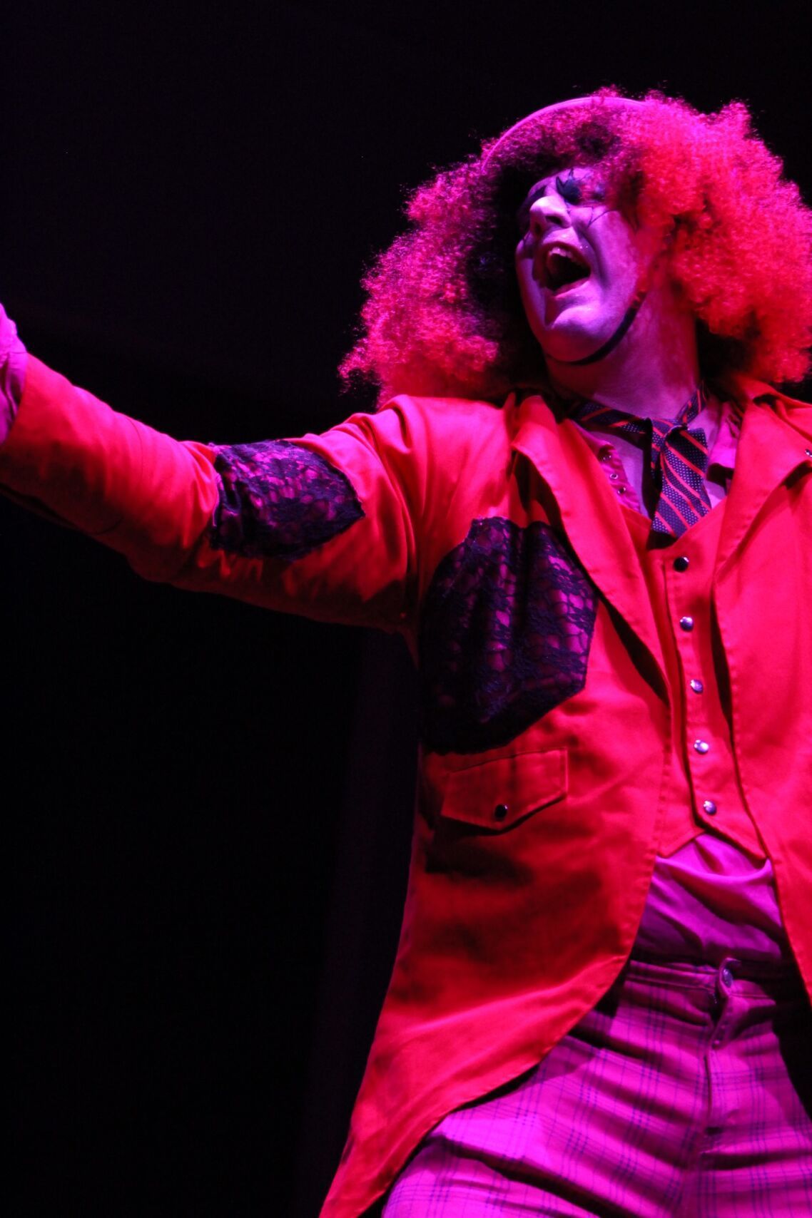 A person with a large red afro and red suit, wide-mouthed, on a dark stage with purple lighting.