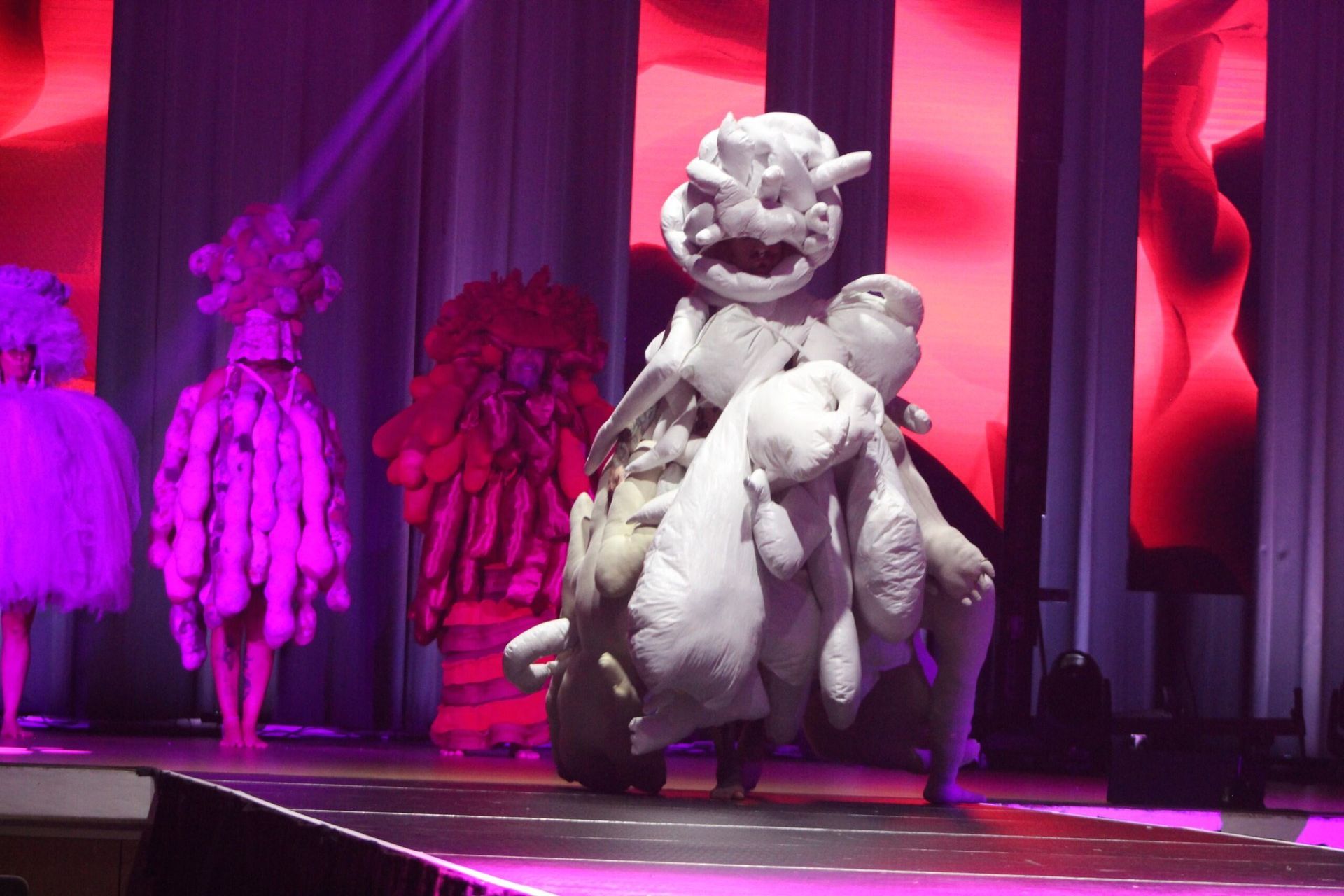 A person in a large, white, textured costume walks on a stage with other costumed figures.