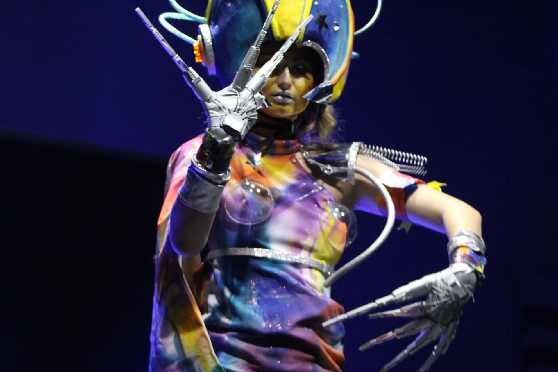 Woman in futuristic costume with clawed hands and glowing headpiece, blue stage backdrop.