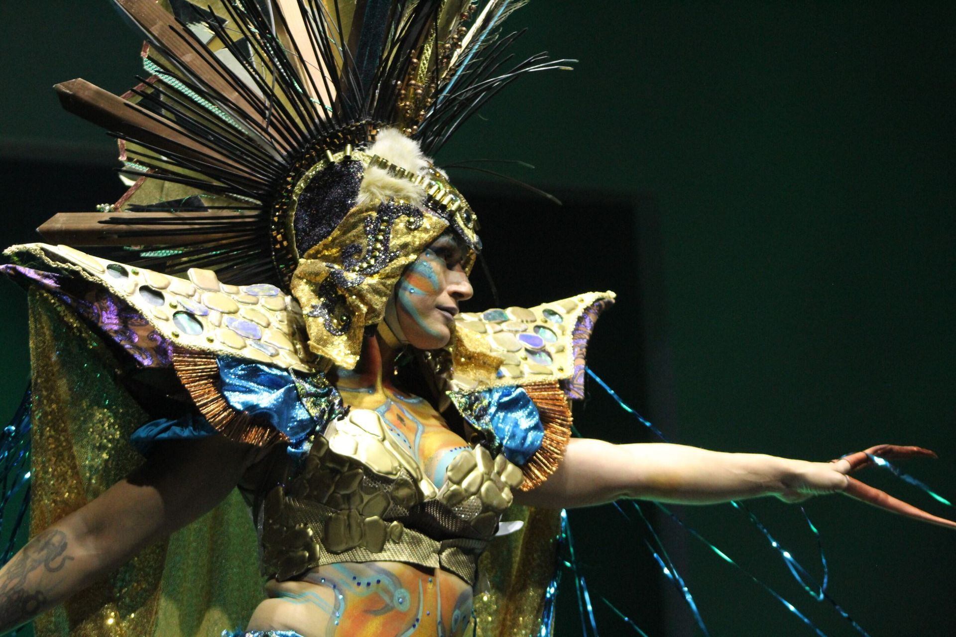 Person in elaborate gold costume, headdress, face paint, and long fingernails, performing onstage.