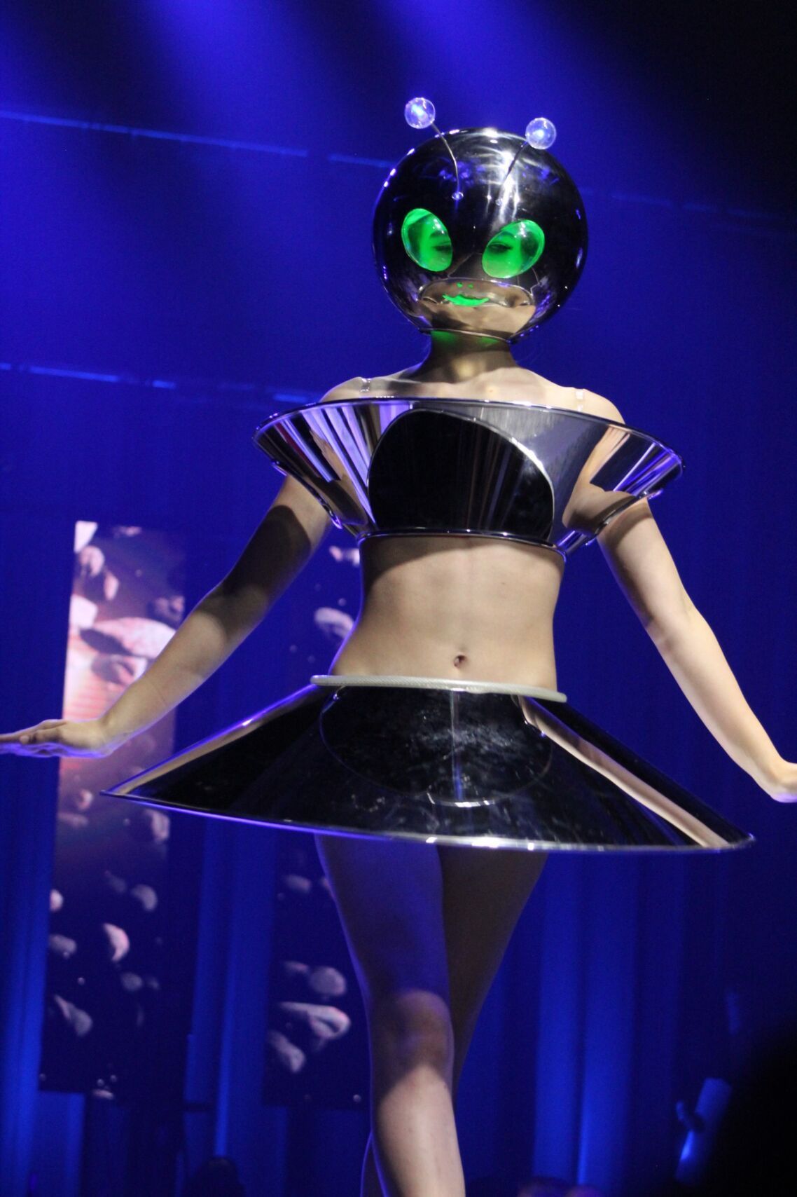 Woman in futuristic alien costume with metallic head and bodywear, posed on stage.
