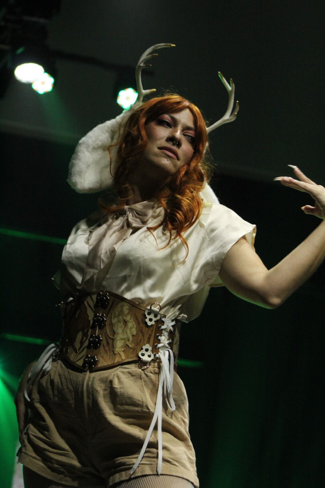 Woman in deer-themed costume, red hair, stage setting, wearing antlers, corset, and shorts, striking a pose.