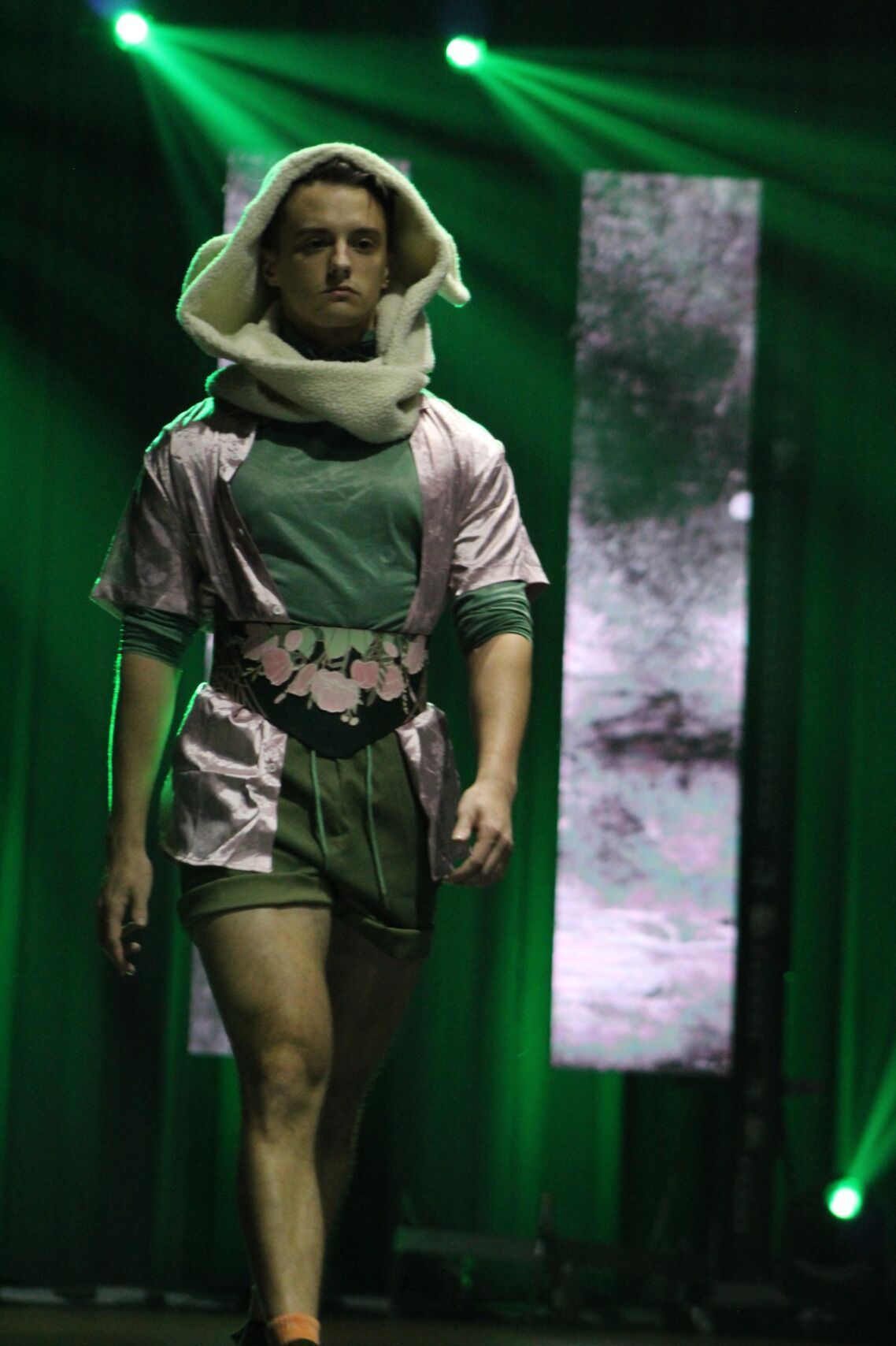 Man in runway fashion; pink top, green shorts, hooded scarf. Green and white stage background.
