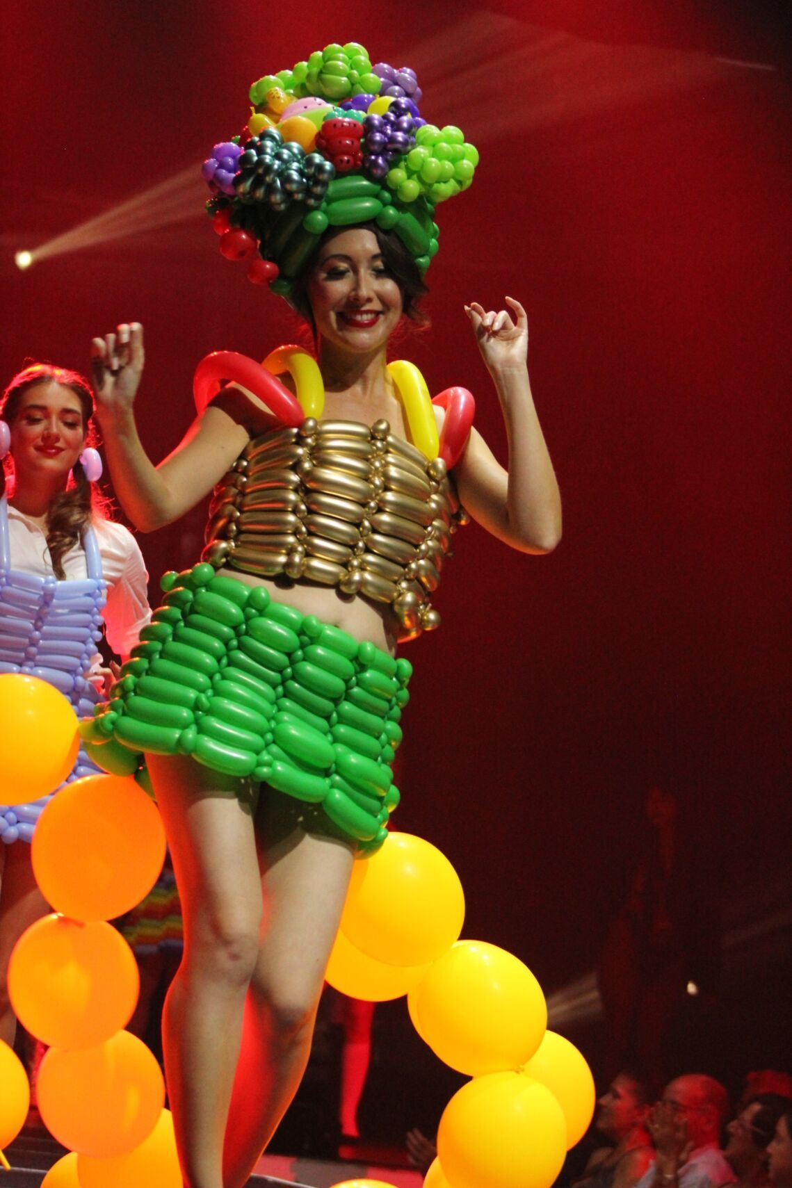 Woman in a balloon dress and headdress, smiling and dancing on a stage with red lighting.