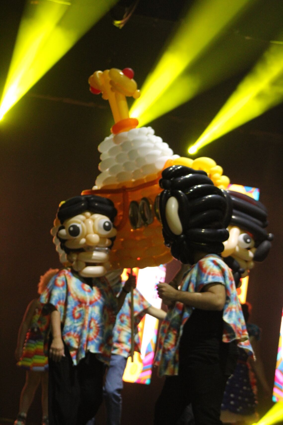 People in masks and Hawaiian shirts holding a large balloon sculpture onstage with yellow spotlights.