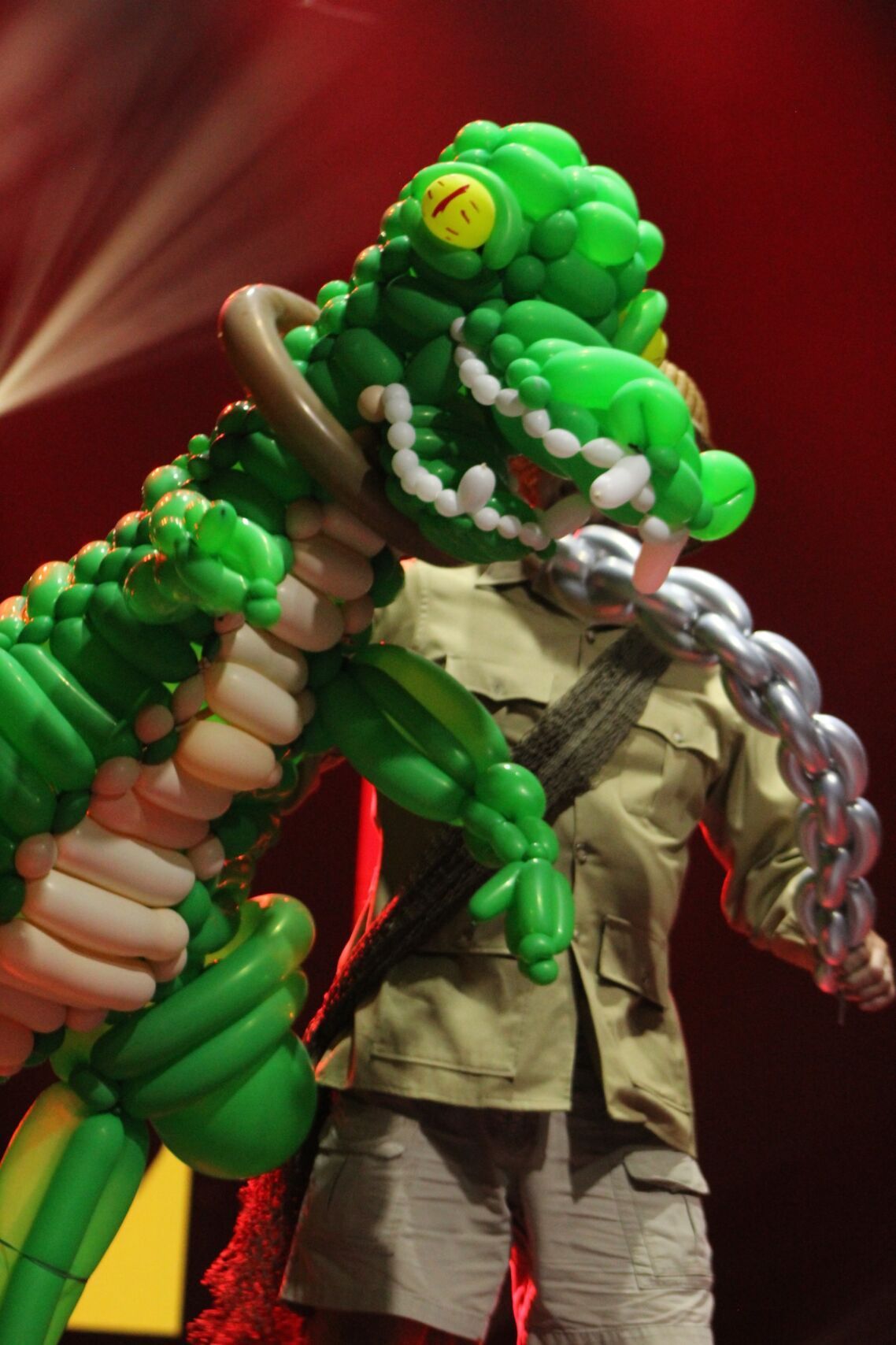 A person in safari clothes holds a large green balloon crocodile, mouth open, chain around its neck, red background.